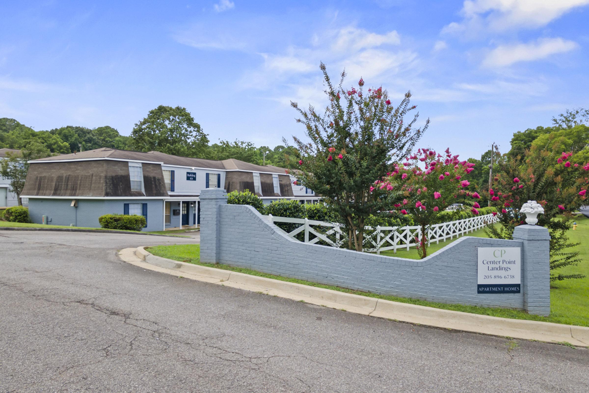 A sign for Center Point Landing, featuring a white fence and flowering bushes. The building in the background is low-rise and has a gray exterior. The scene is set in a green, landscaped area under a clear blue sky.