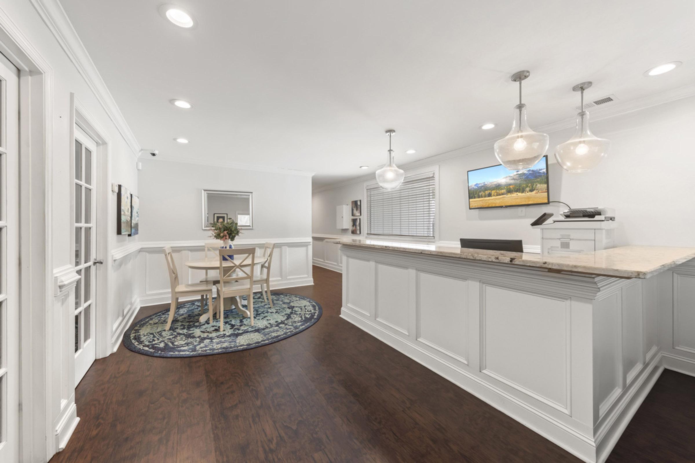 A modern, well-lit reception area featuring a wooden floor, elegant white walls, and a small round table with flowers. A large rug adds color, and pendant lighting hangs above the reception desk, which has a computer and a flat-screen TV mounted on the wall.