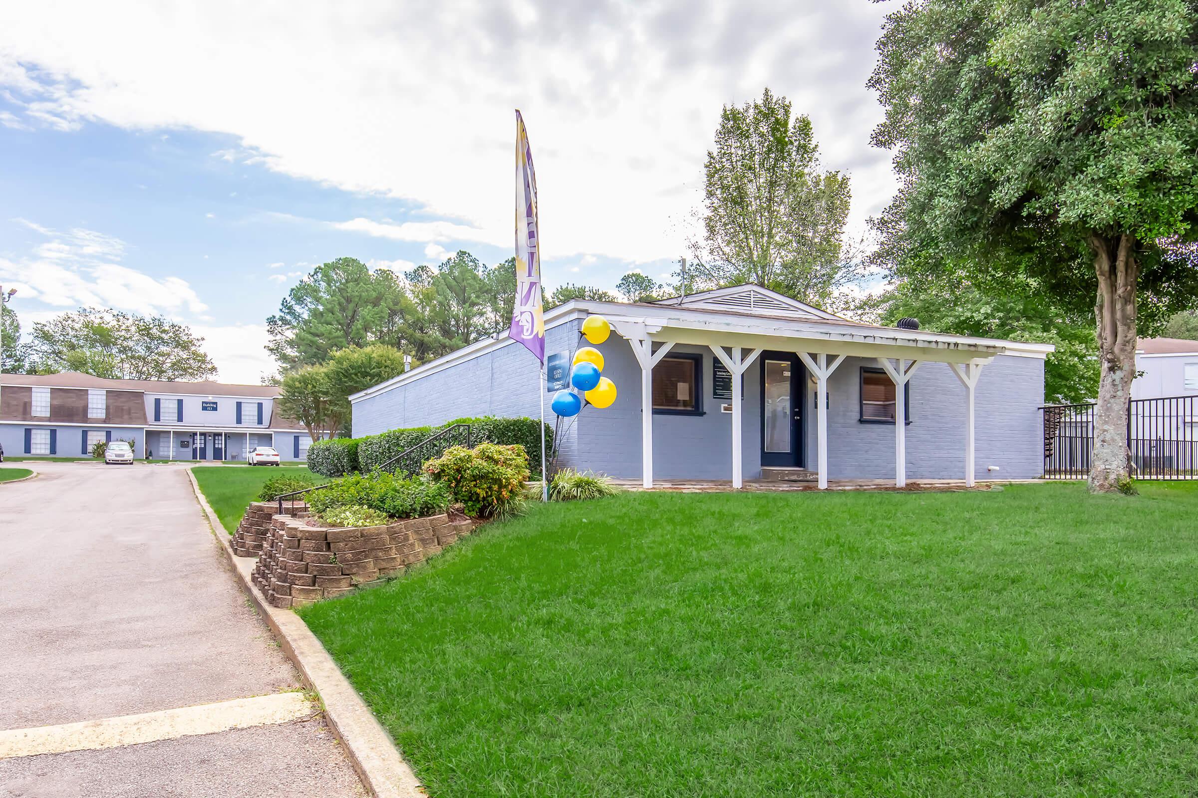 A single-story blue house with a landscaped lawn, featuring a canopy entrance and colorful balloons. In the background, there are tree-lined streets and additional residential buildings. The sky is partly cloudy, adding a pleasant atmosphere to the scene.
