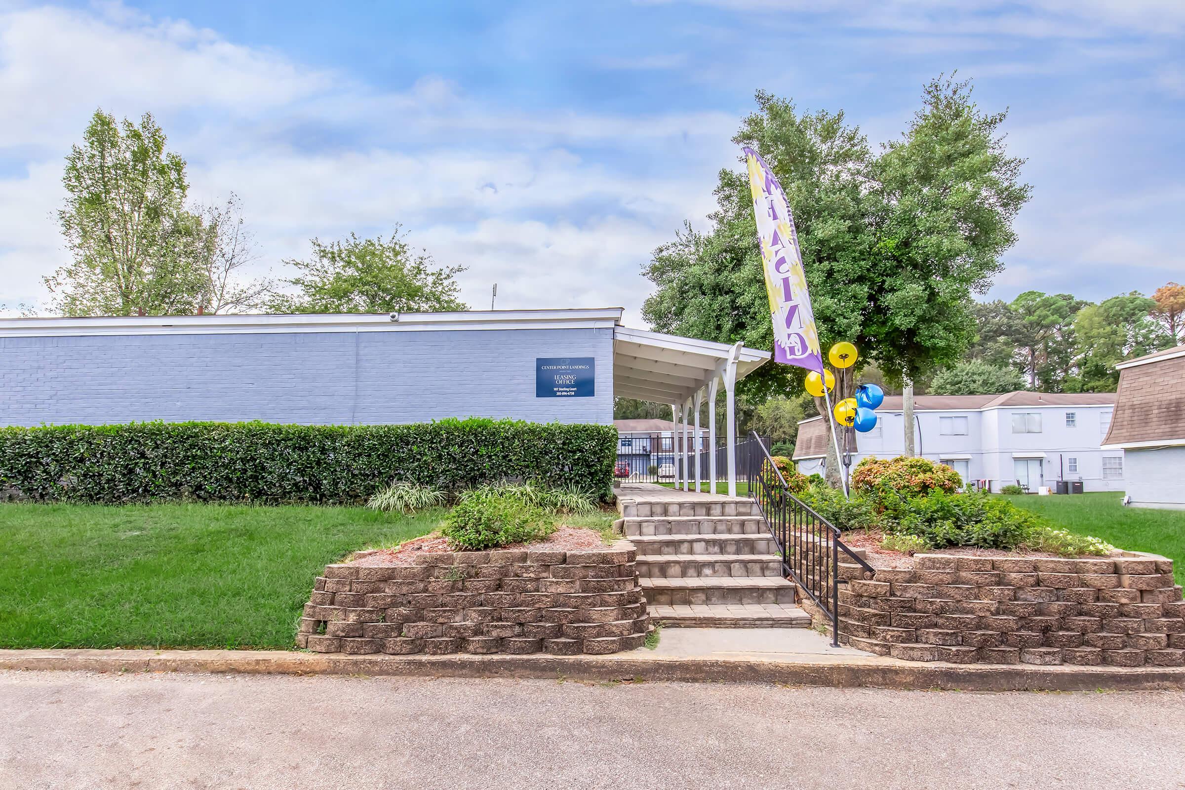 Exterior view of a gray building with a welcoming entrance featuring steps and a ramp. The area is landscaped with green grass and shrubs. A colorful flag and balloons are displayed, indicating an event or celebration. Trees are visible in the background under a partly cloudy sky.