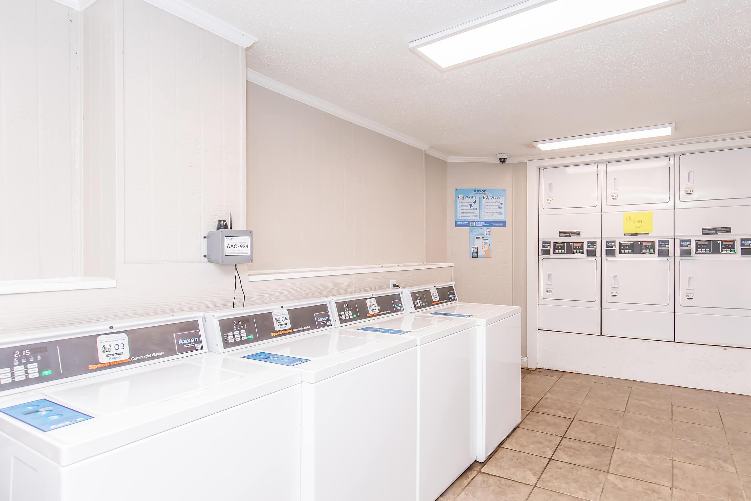 A clean, brightly lit laundry room featuring several white washing machines and dryers, with digital controls. The walls are painted in a light color, and there are utility notices and informational posters on display. The floor is tiled, and the overall space appears well-maintained and organized.