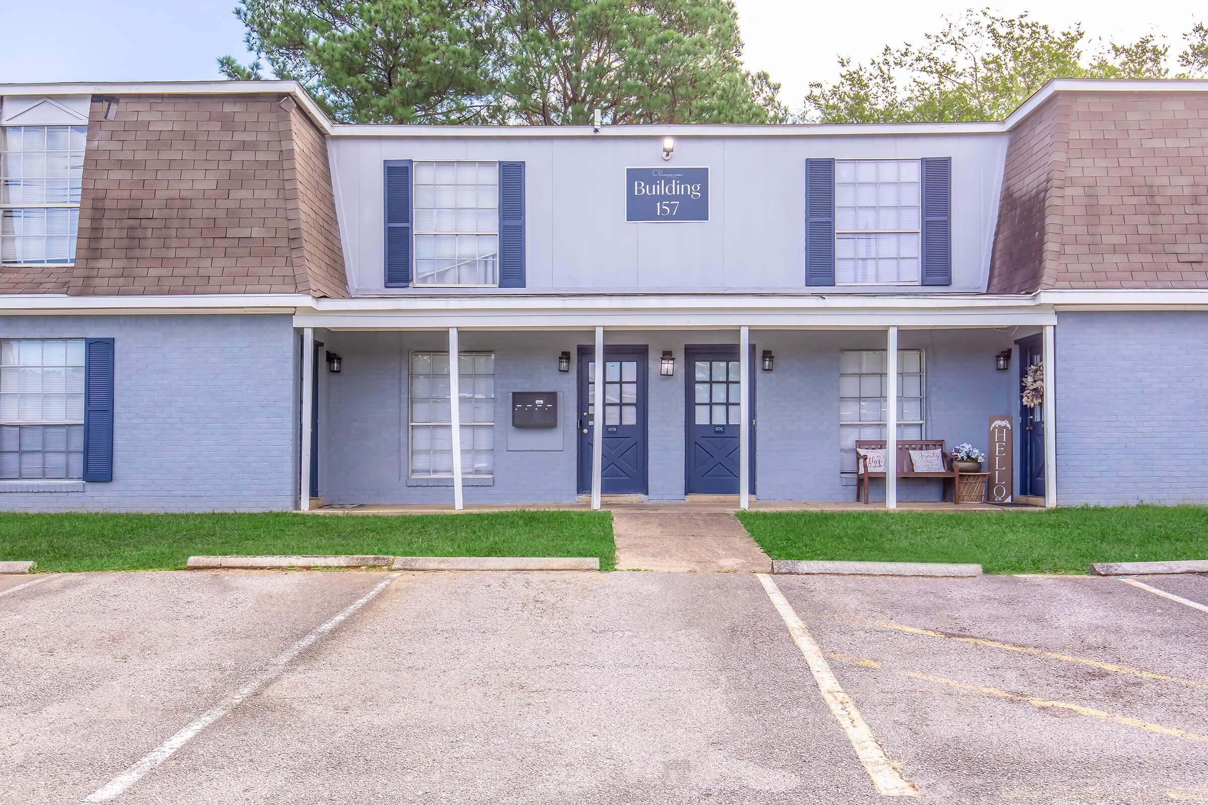 Exterior view of a two-story building labeled "Building 157." The façade features a light blue color, dark blue window shutters, and a sloped brown roof. The entrance has double doors, flanked by large windows and outdoor seating. The surrounding area includes a parking lot and grassy lawn.