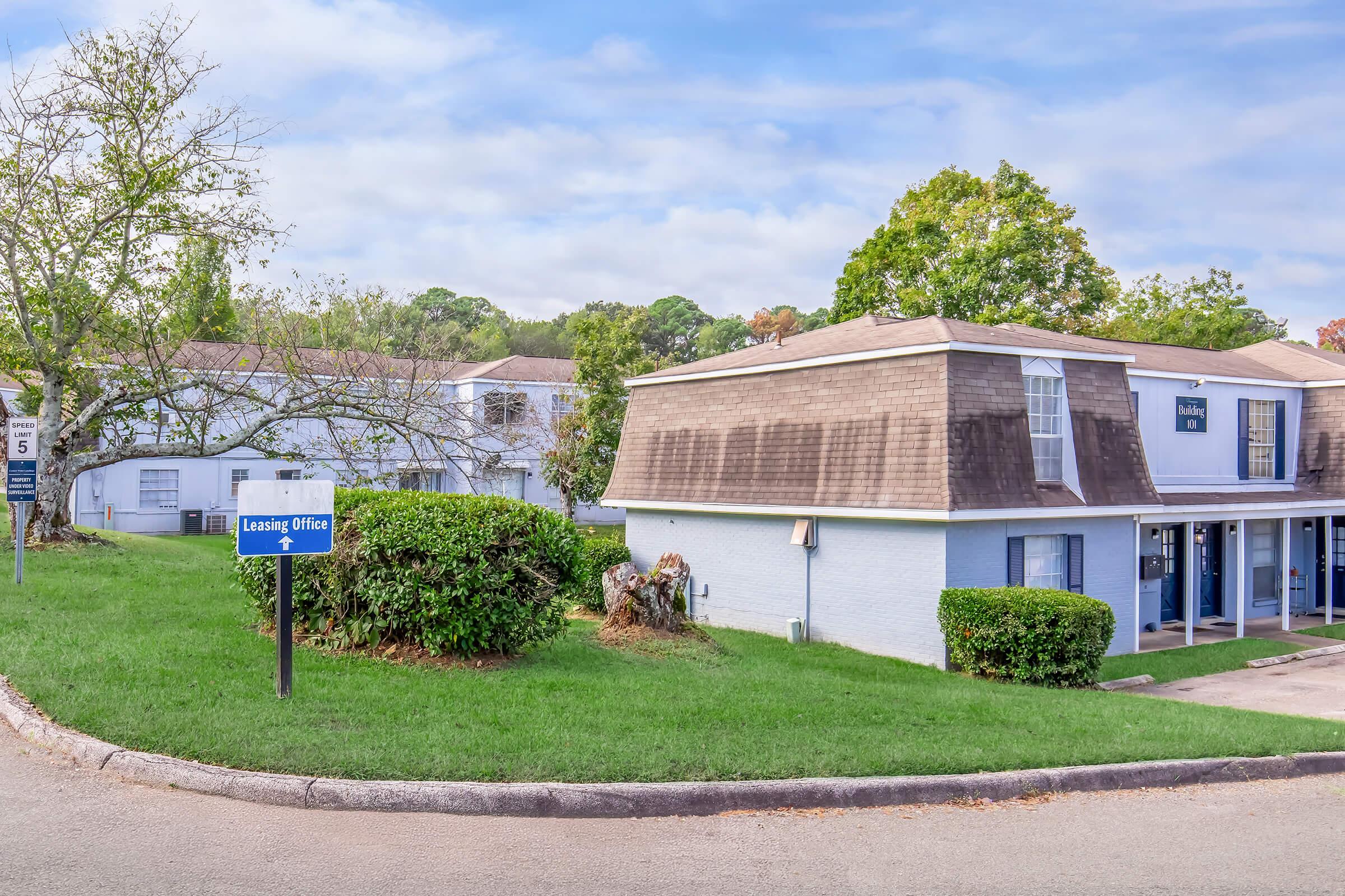 A leasing office building in a residential complex, featuring a sign indicating "Leasing Office." The architecture includes a sloped roof and adjacent greenery. The surrounding area has grass and a few trees, with a clear blue sky in the background.