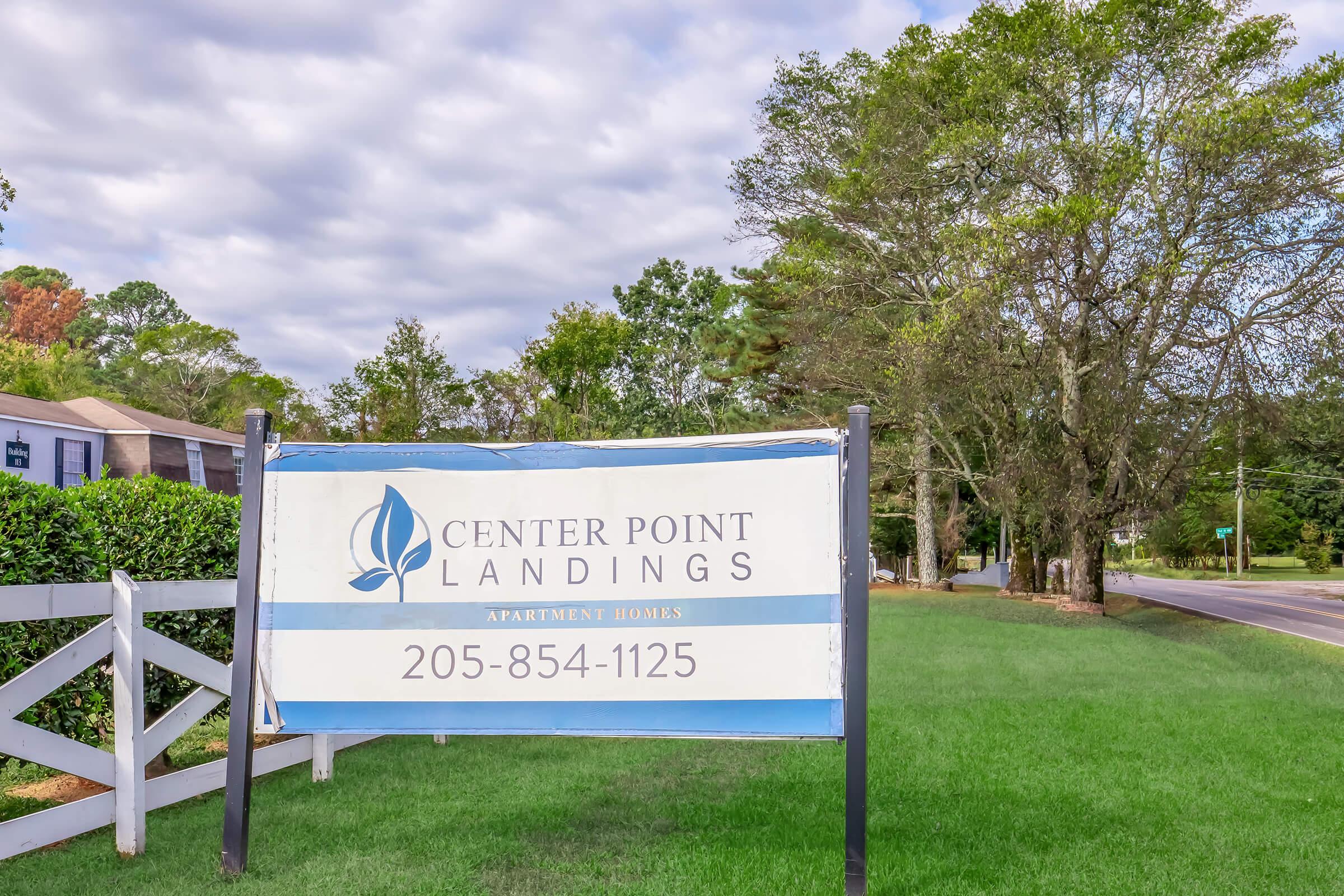 Sign for Center Point Landings Apartment Homes featuring the name and contact number 205-854-1125, positioned in a grassy area with trees in the background and a cloudy sky above.