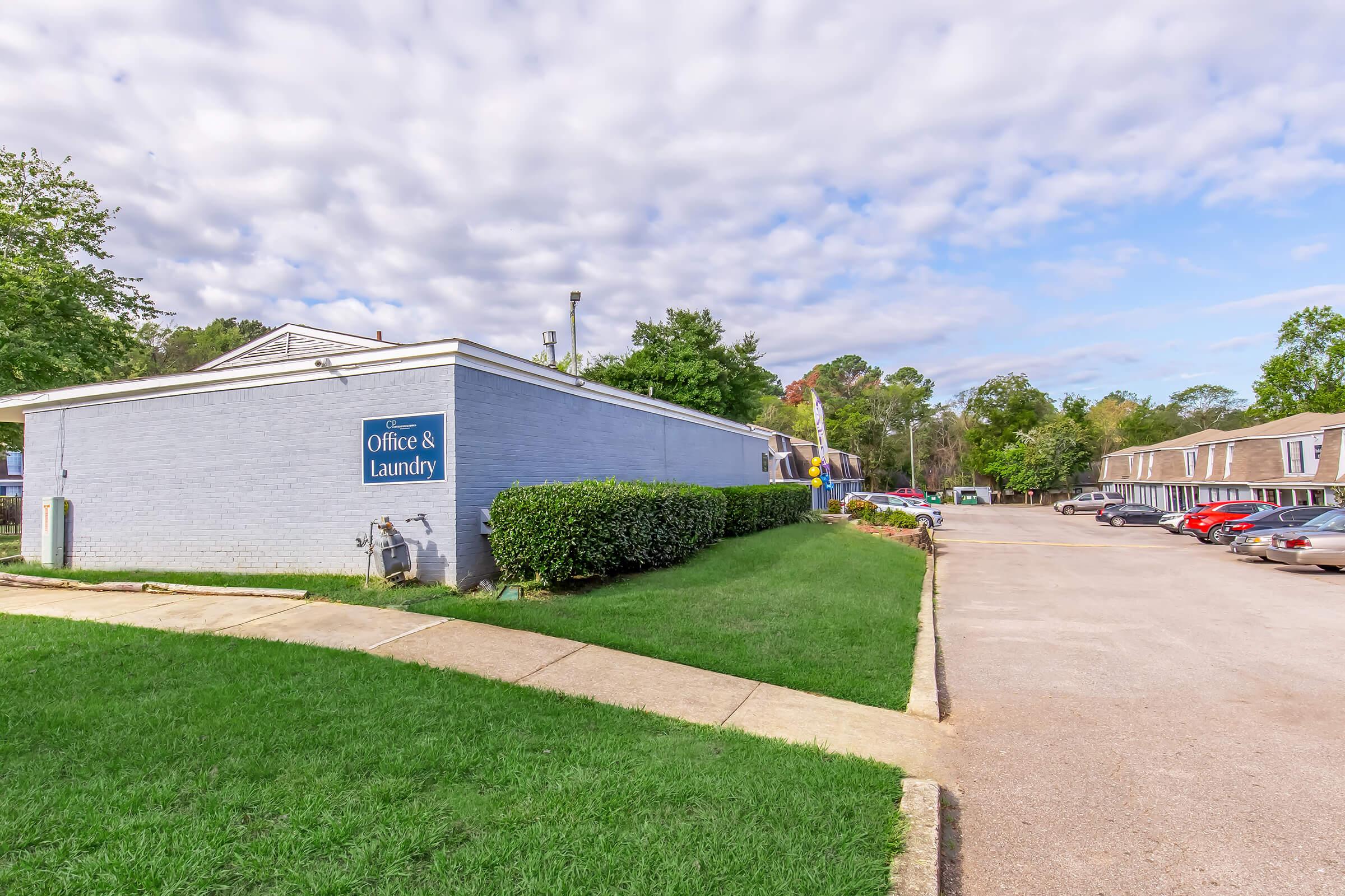 A gray building with a blue sign reading "Office & Laundry" is situated on the left. The area is well-maintained with green grass and shrubs. In the background, a row of vehicles is parked along a gravel pathway, and there are trees lining the background under a partly cloudy sky.