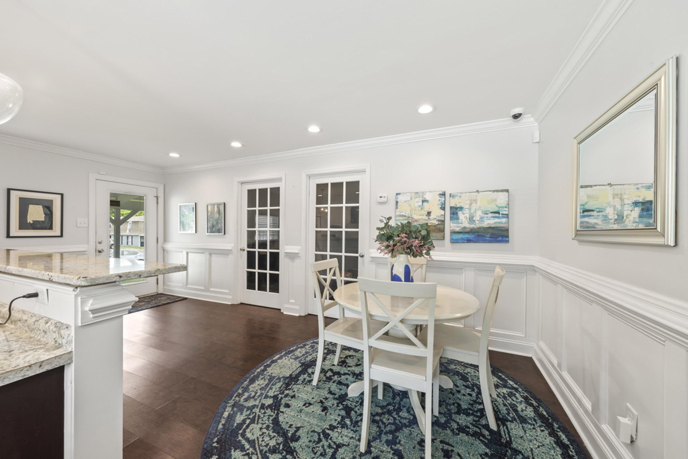 A bright, modern dining area featuring a round table with white chairs, surrounded by light-colored walls and wainscoting. Framed artwork adorns the walls, and a large area rug adds color to the hardwood floor. French doors lead to the outside, enhancing the room's spacious feel.