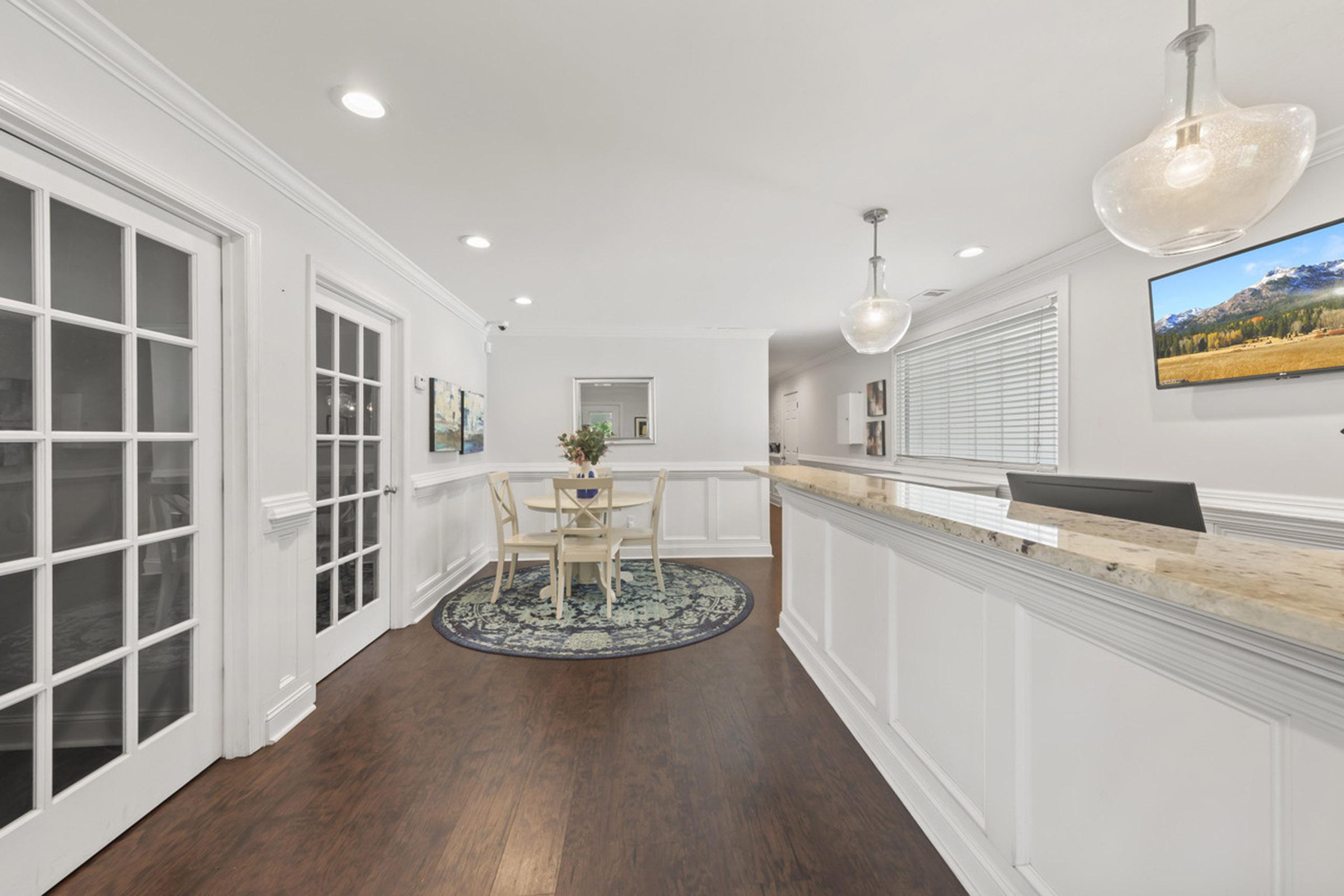 A modern reception area featuring a white and wood counter, pendant lighting, and a small round table with chairs. A colorful rug lies on the dark hardwood floor, and large windows provide natural light. Images of landscapes hang on the walls. The atmosphere is bright and welcoming.