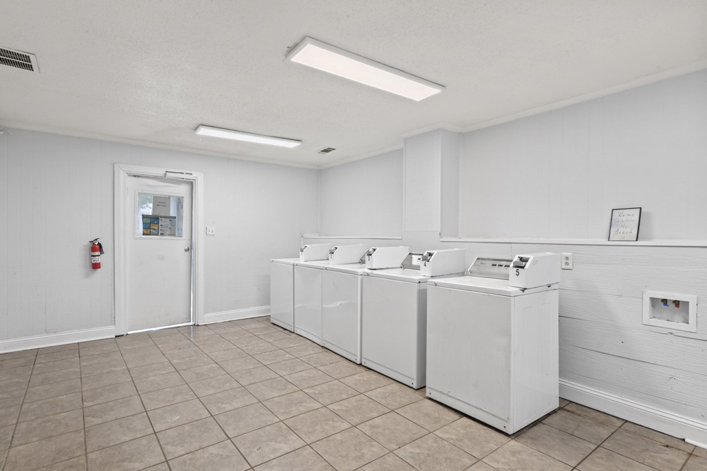 A clean, well-lit laundry room featuring four white washing machines aligned against a wall, with beige tiled flooring. There is a door leading outside, a small framed picture on the wall, and a fire extinguisher mounted near the door. The walls are painted a light color, enhancing the bright and spacious atmosphere.