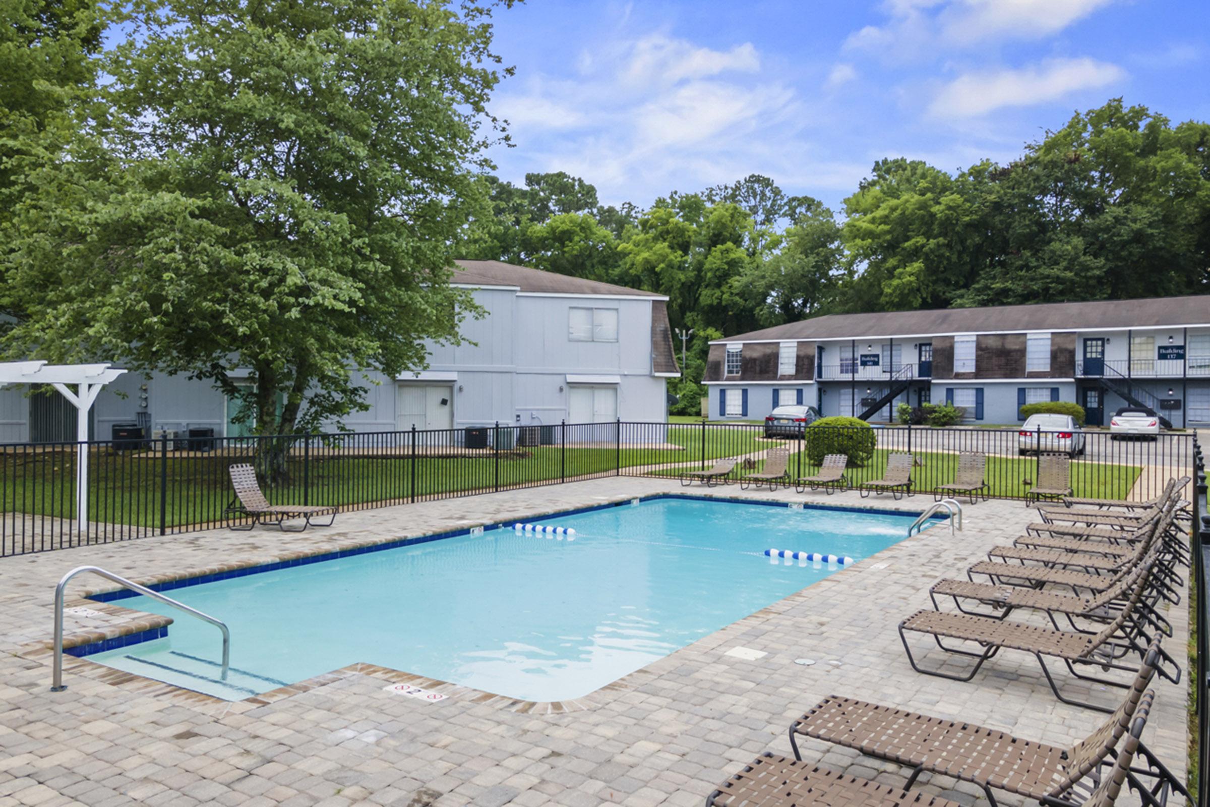 A clear blue swimming pool surrounded by lounge chairs, with a manicured lawn and trees in the background. Nearby buildings reflect a residential style, featuring multiple units, and a parking area is visible in the distance. The scene is bright and inviting, indicating a well-maintained outdoor space.