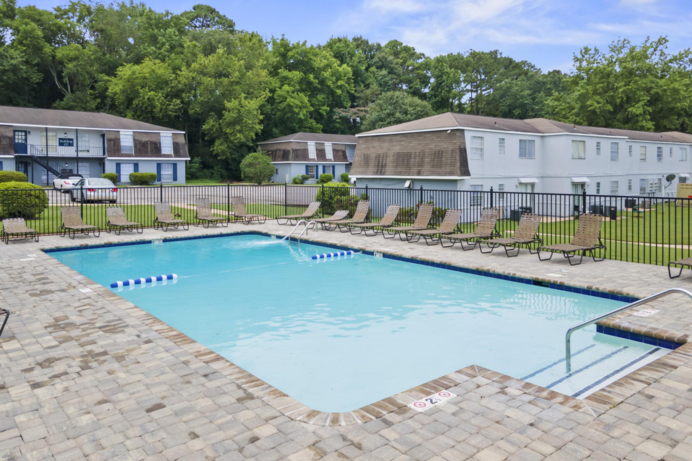 A clear swimming pool surrounded by lounge chairs, located in a landscaped area with trees and two multi-story apartment buildings in the background. The pool area features a stone deck and safety lane markers. The scene is inviting and well-maintained.