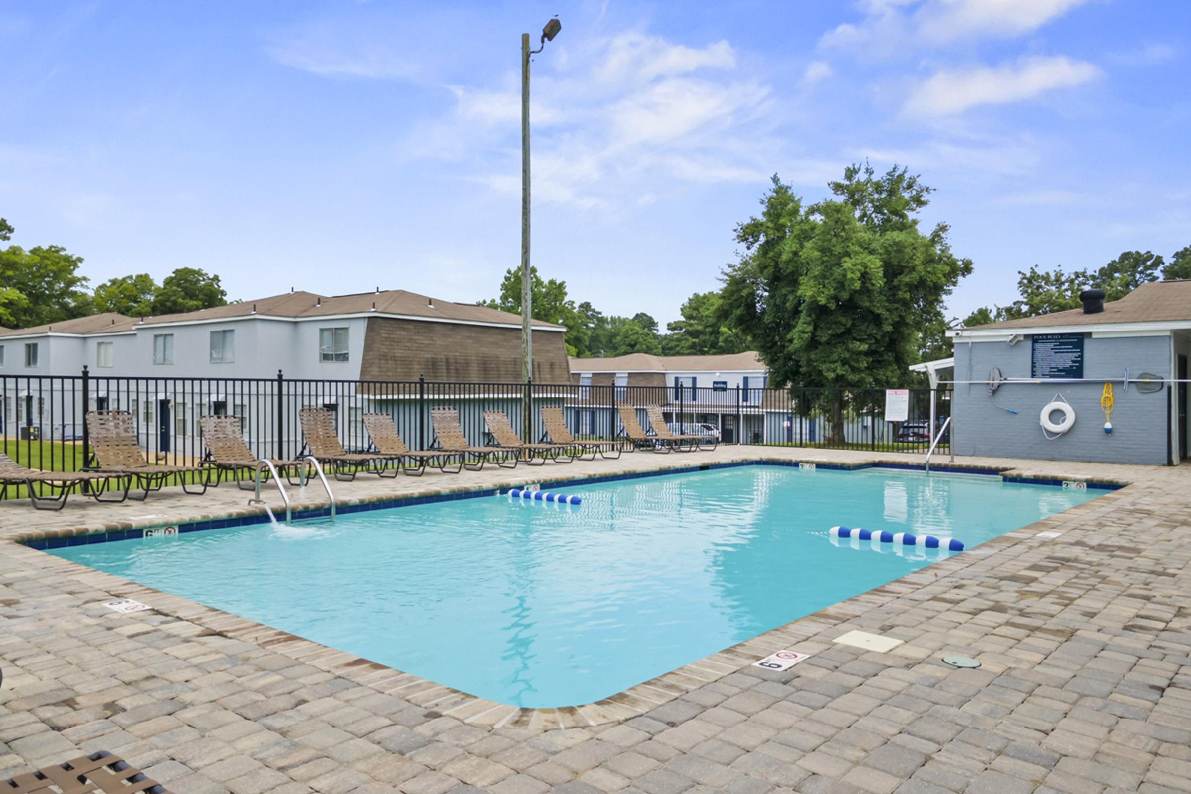 A clean, inviting outdoor swimming pool surrounded by lounge chairs and a fence. The water is bright blue, and there are float markers in the pool. Nearby, there are trees and buildings, with a clear sky overhead, creating a relaxing atmosphere for leisure and recreation.
