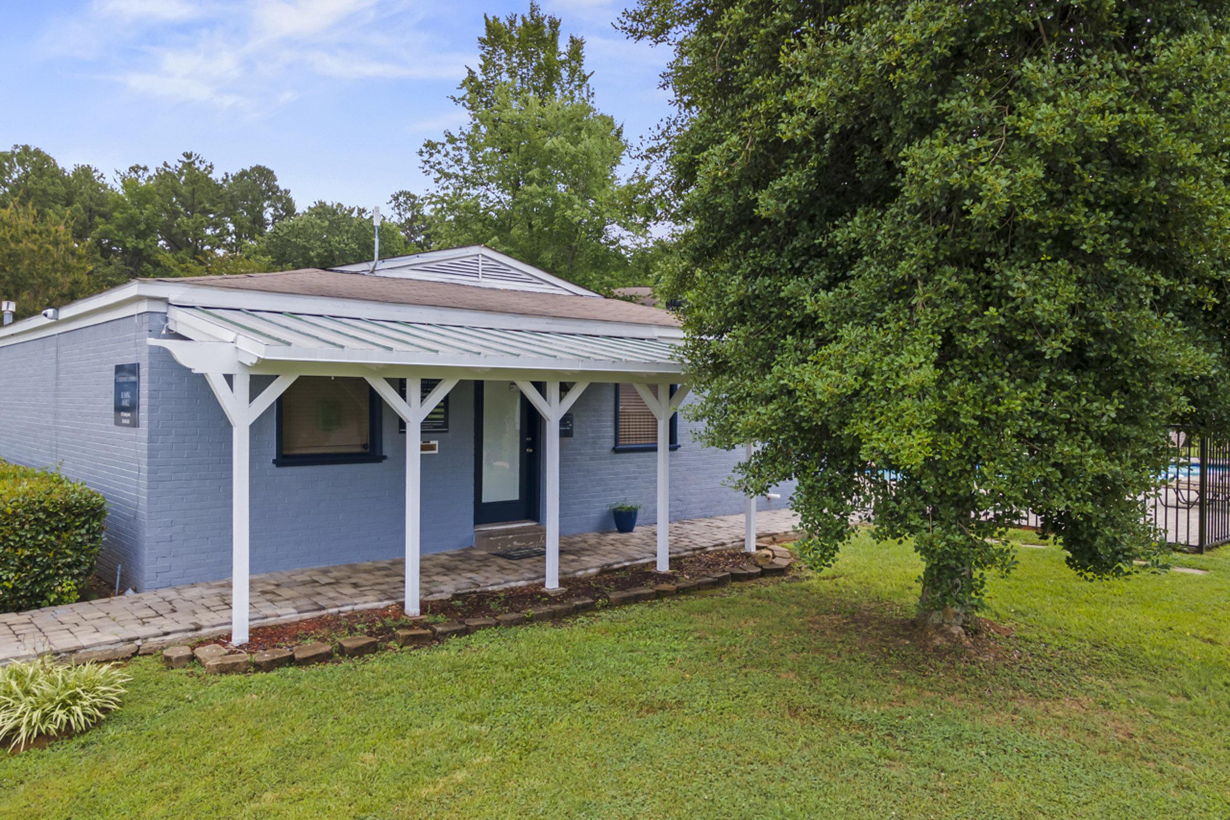 A blue-painted house with a front porch, surrounded by green grass and shrubs. The porch has white columns, and there's a small potted plant at the entrance. In the background, trees can be seen, and a swimming pool is partially visible beyond the house. The sky is clear with a few clouds.