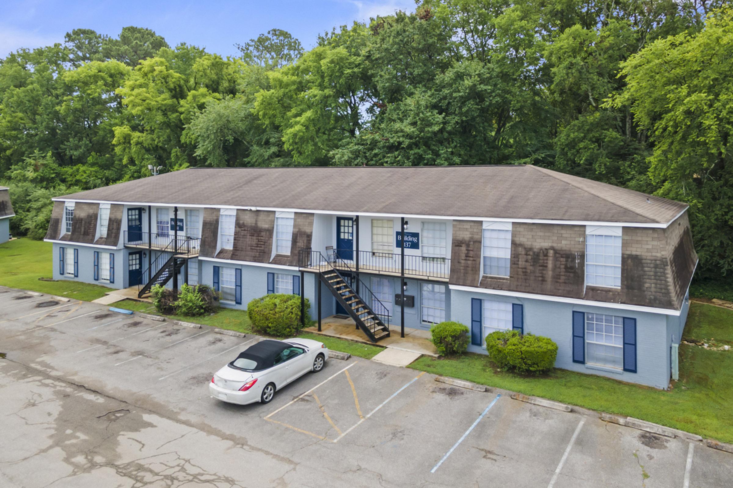 A two-story apartment building with a light blue exterior and brown shingle roofing. There are several windows on each side, and black staircases leading to the second floor. In the foreground, a parked white car is visible in a partially paved parking lot, surrounded by greenery and trees.