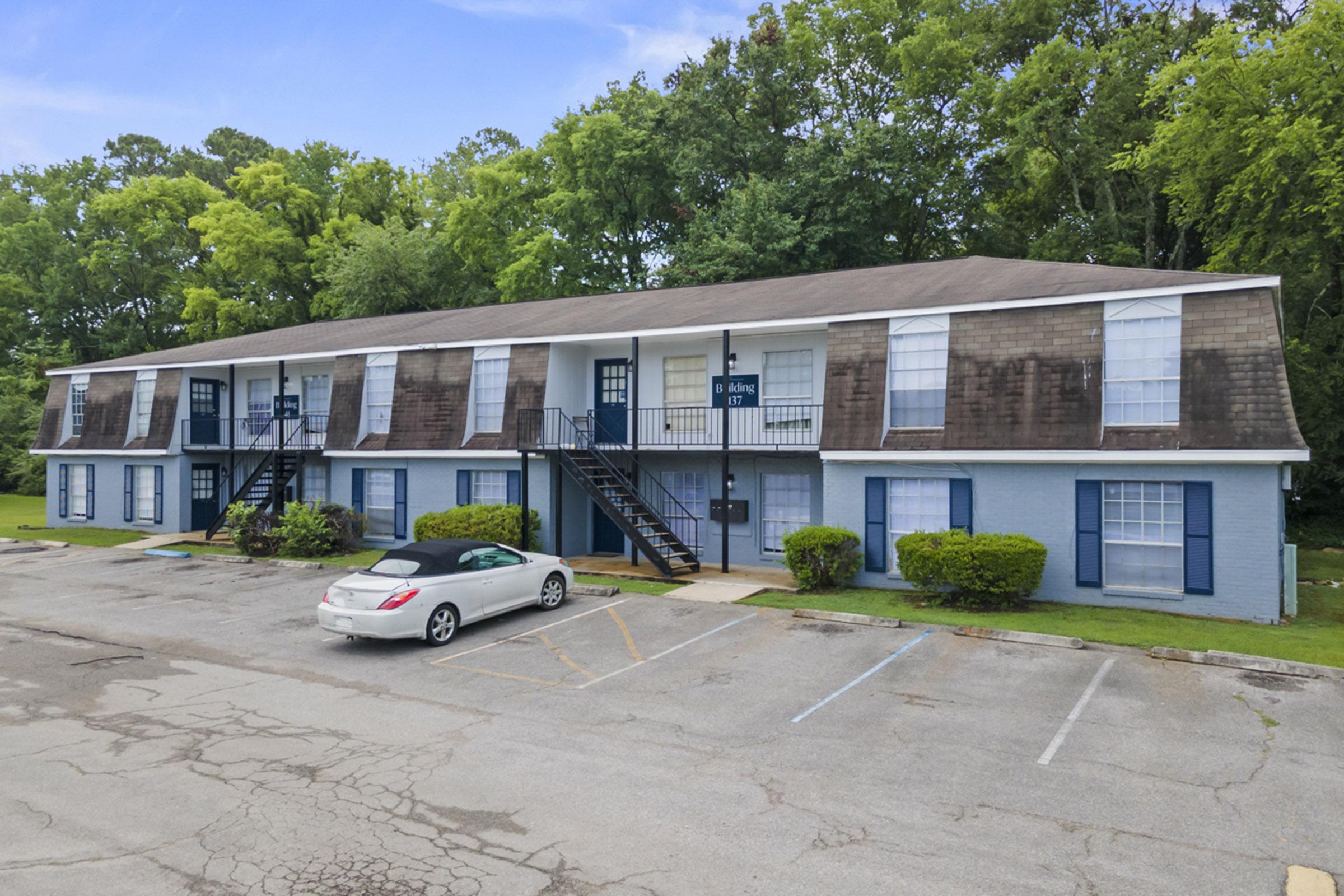 A two-story apartment building with blue siding and white shutters, surrounded by green trees. The building has staircases leading to the upper level, and several parking spaces in front. The area appears well-maintained, with grass and shrubs lining the front of the property.