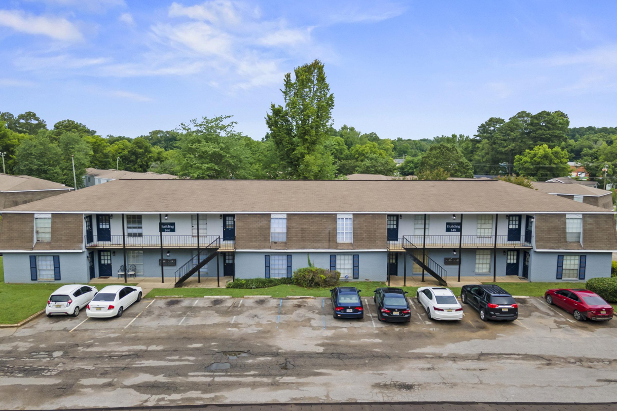 A view of a low-rise apartment complex with two sections, featuring multiple entrances and a parking lot in front. Several cars are parked in the lot, and there are trees and greenery in the background, under a clear blue sky. The structure has a light brown exterior with blue doors and windows.