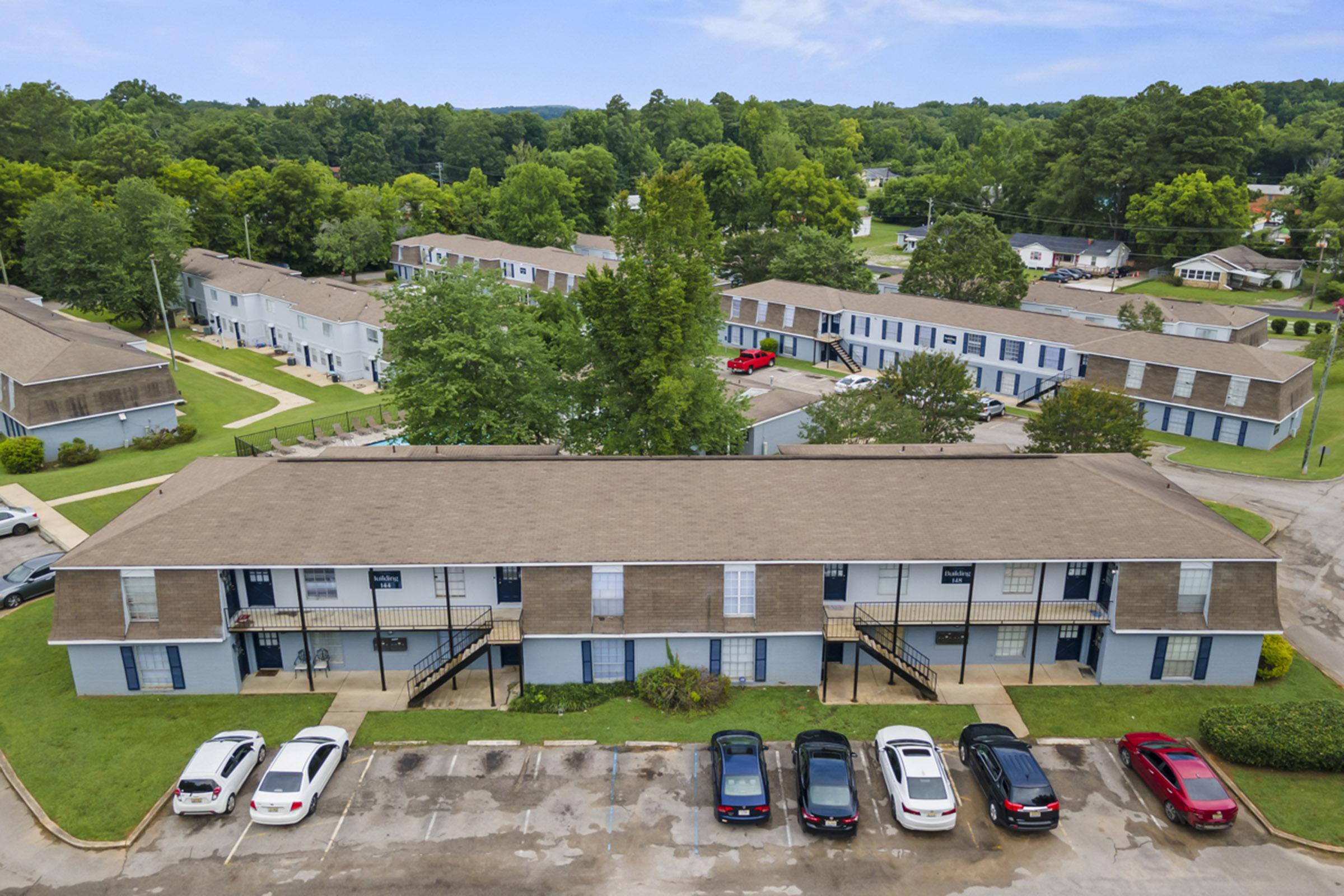 Aerial view of a residential complex featuring two-story apartment buildings with blue shutters. The surrounding area includes green trees and a parking lot with several cars. The complex has manicured lawns and walkways, situated in a suburban setting.