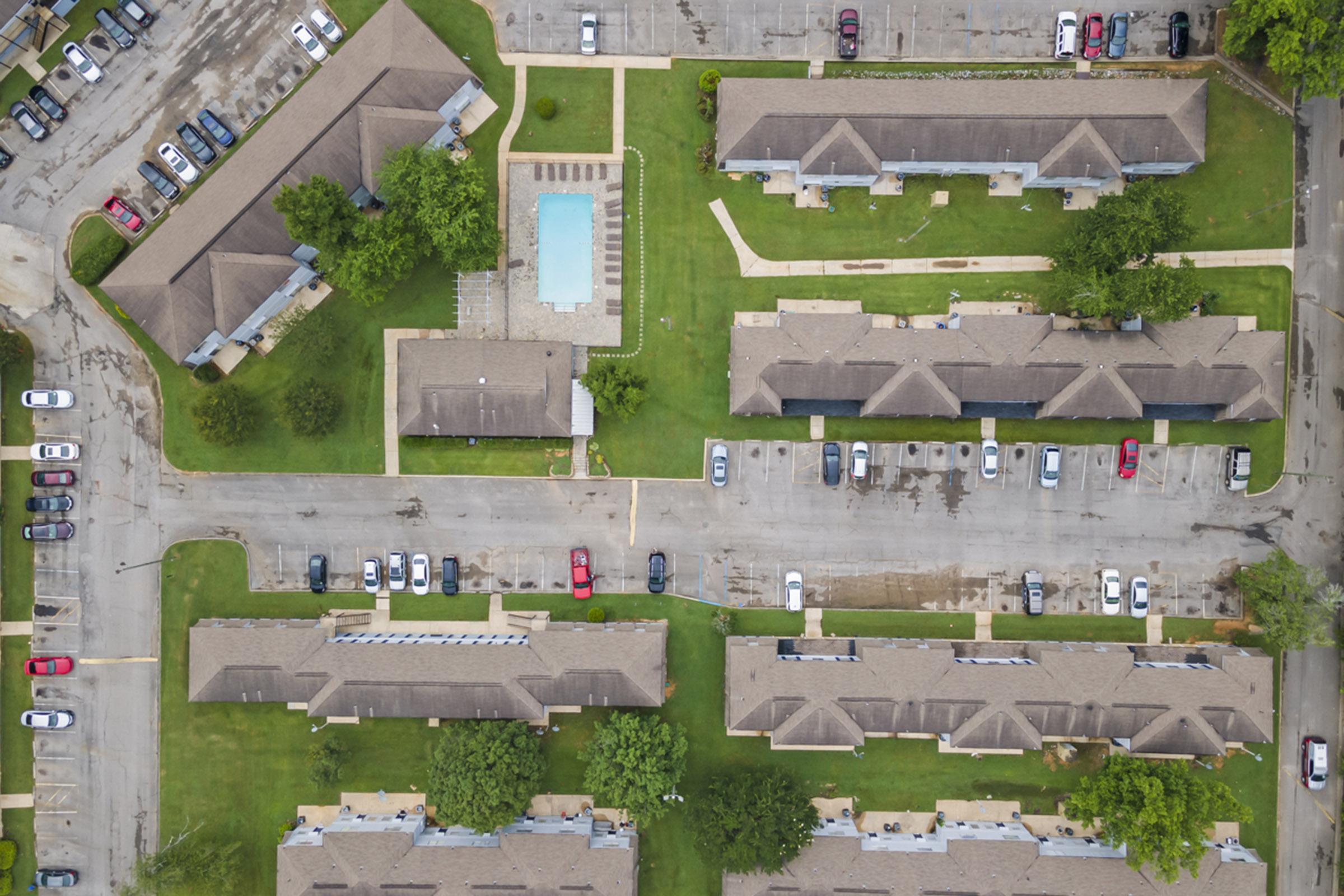 Aerial view of a residential complex featuring multiple single-story buildings, a pool area with lounge chairs, and surrounding green lawns. Nearby, there are parked cars along the streets and pathways connecting the buildings.