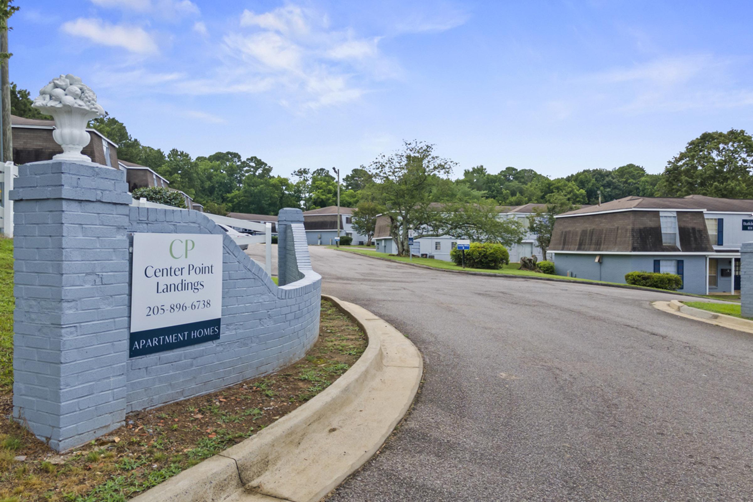 A view of the entrance to Center Point Landings, a quiet apartment complex. The sign displays "CP Center Point Landings" along with a phone number. The road leads into the complex surrounded by green trees and well-maintained landscaping, giving a serene suburban atmosphere.