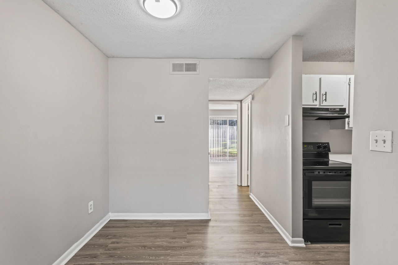 A view of a small, light-colored hallway leading to an open kitchen area. The walls are painted light gray, and the floor features brown vinyl planks. A thermostat is mounted on the wall, and there is a doorway leading to the living area with large windows visible in the background.