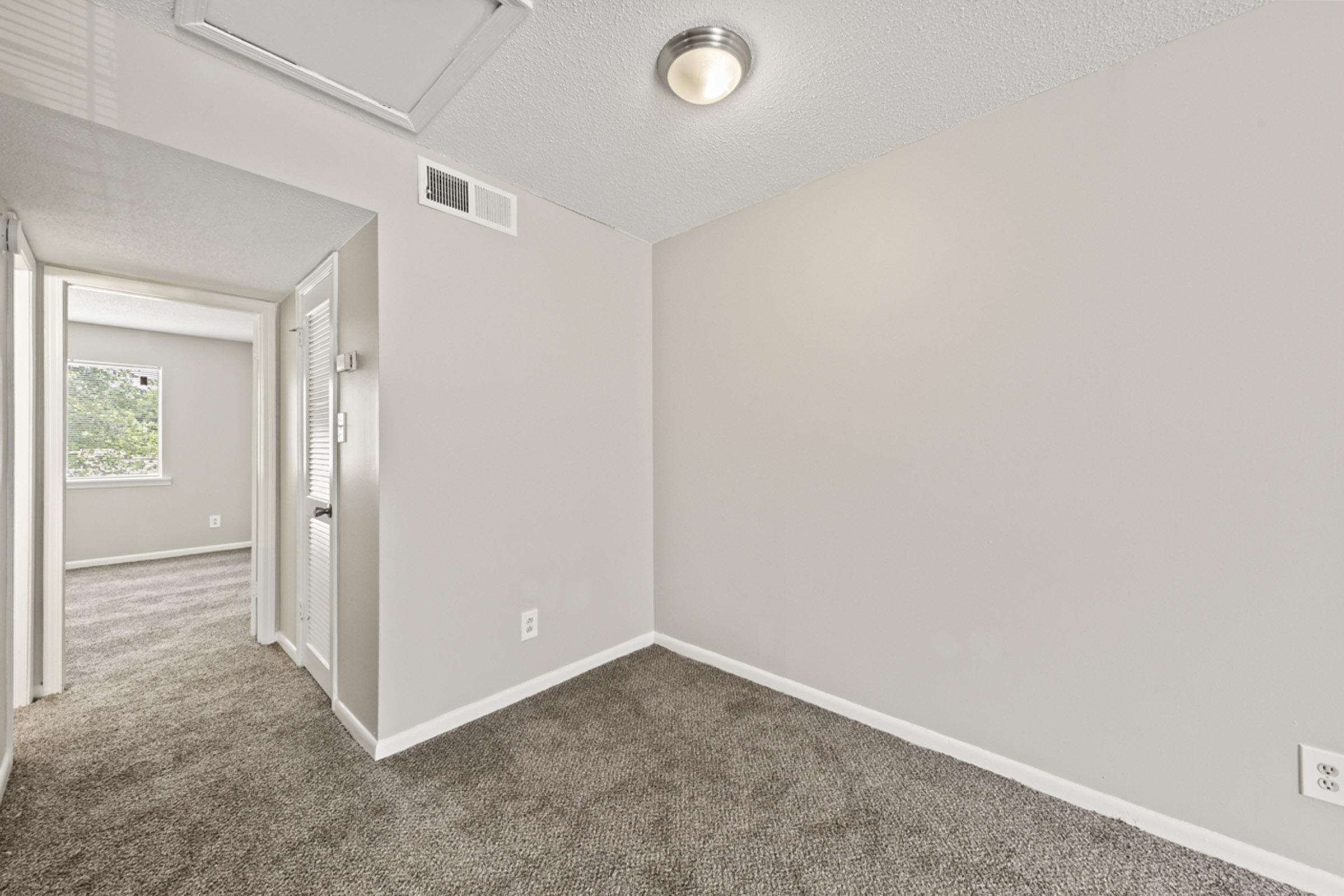 A well-lit room featuring beige walls and carpeted flooring. There is a ceiling light fixture and a window partially visible in the background. The room appears empty, with a doorway leading to another area, giving a sense of spaciousness and modern design.