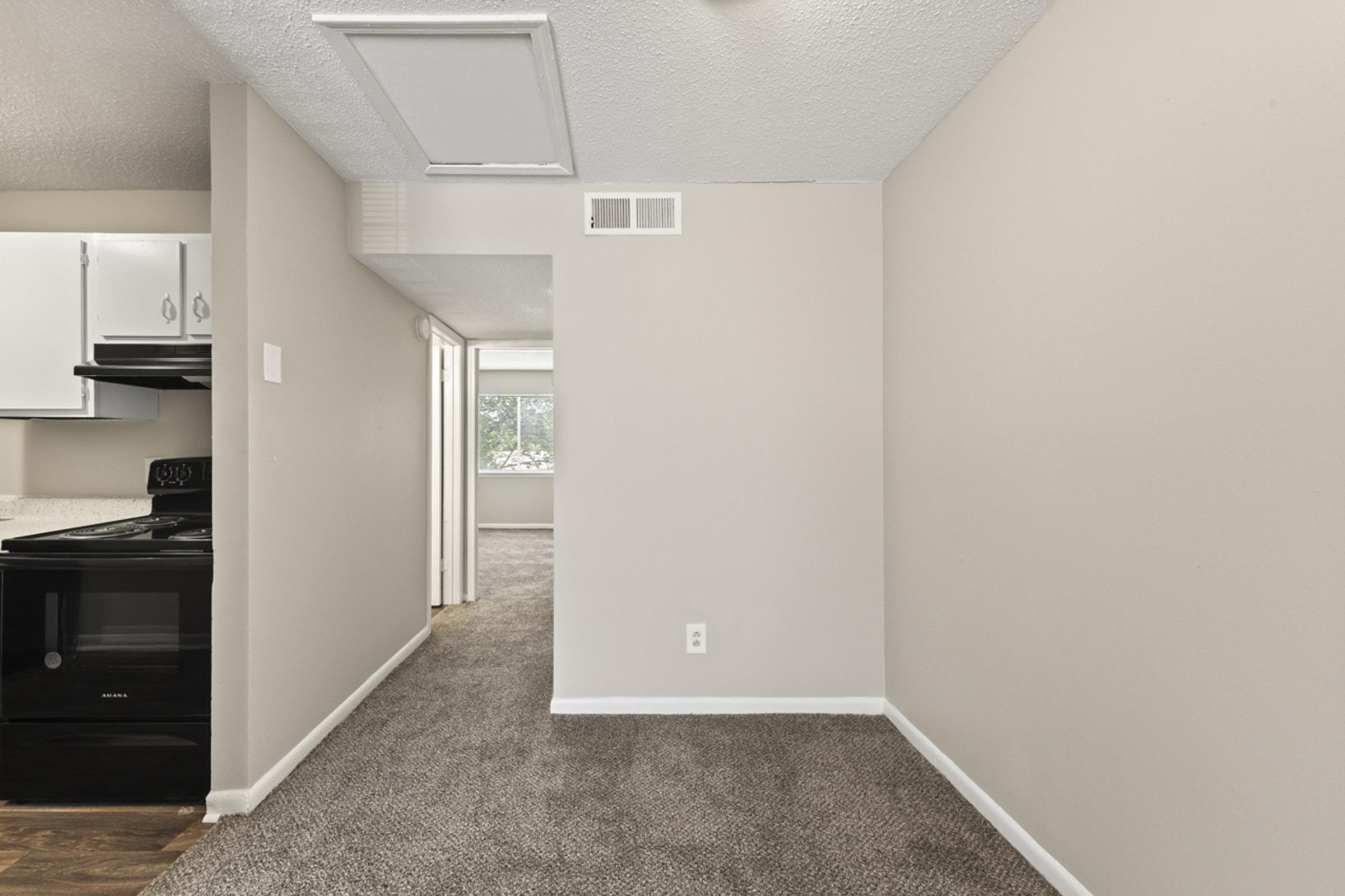 Interior view of an apartment featuring a carpeted living area, light-colored walls, and a doorway leading to additional rooms. A black stove is visible in the kitchen area on the left, and a window can be seen at the end of the hallway, providing natural light.