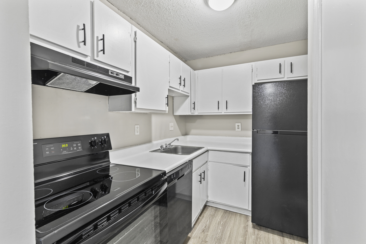 A modern kitchen featuring white cabinets, a black oven and stovetop, a stainless steel sink, and a black refrigerator. The walls are light-colored, and the flooring is a wood-like laminate. The layout is compact and functional, designed for efficient use of space.