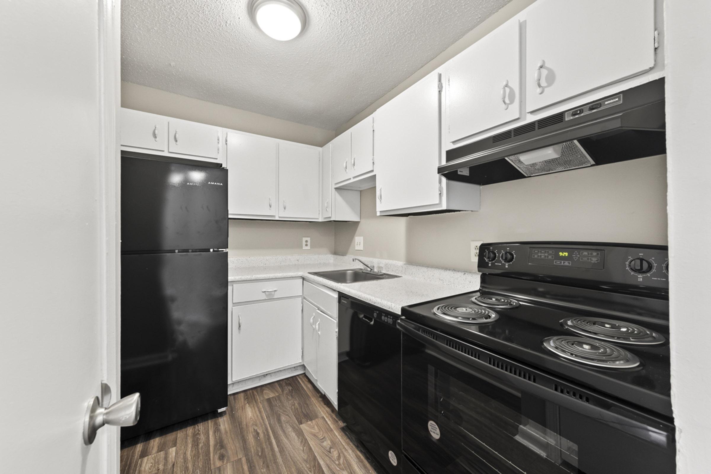 A small, modern kitchen featuring white cabinets, black refrigerator, and an electric stove with an oven. It has a stainless steel sink and granite-like countertops, with wooden flooring. The room is well-lit by a ceiling light, providing a clean and bright cooking space.