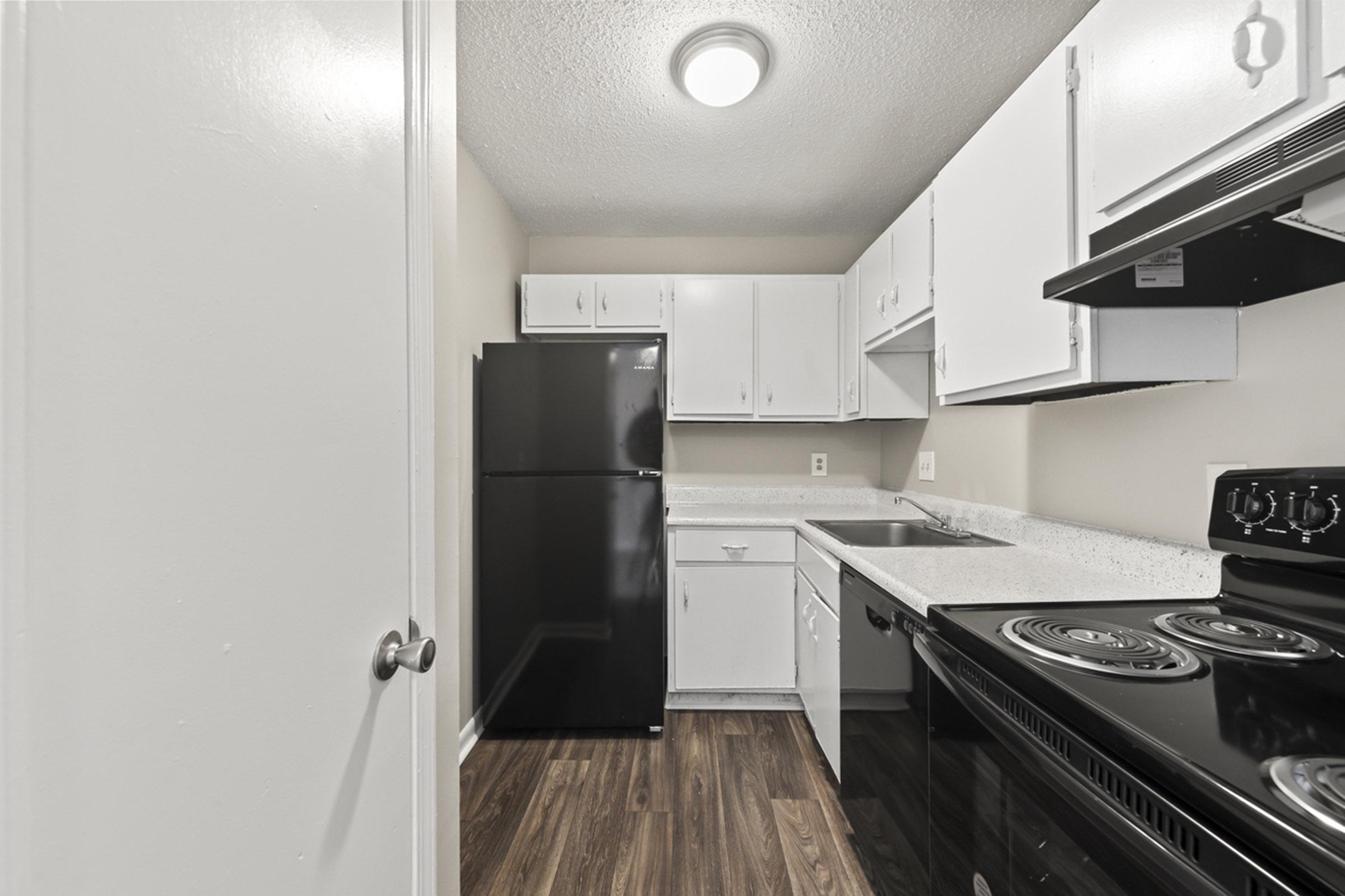 A small kitchen featuring white cabinets, a black refrigerator, and a black stove with an oven. The countertops are light-colored, and the floor is dark wood. A door is visible on the left side, and there is ample overhead lighting in the space.