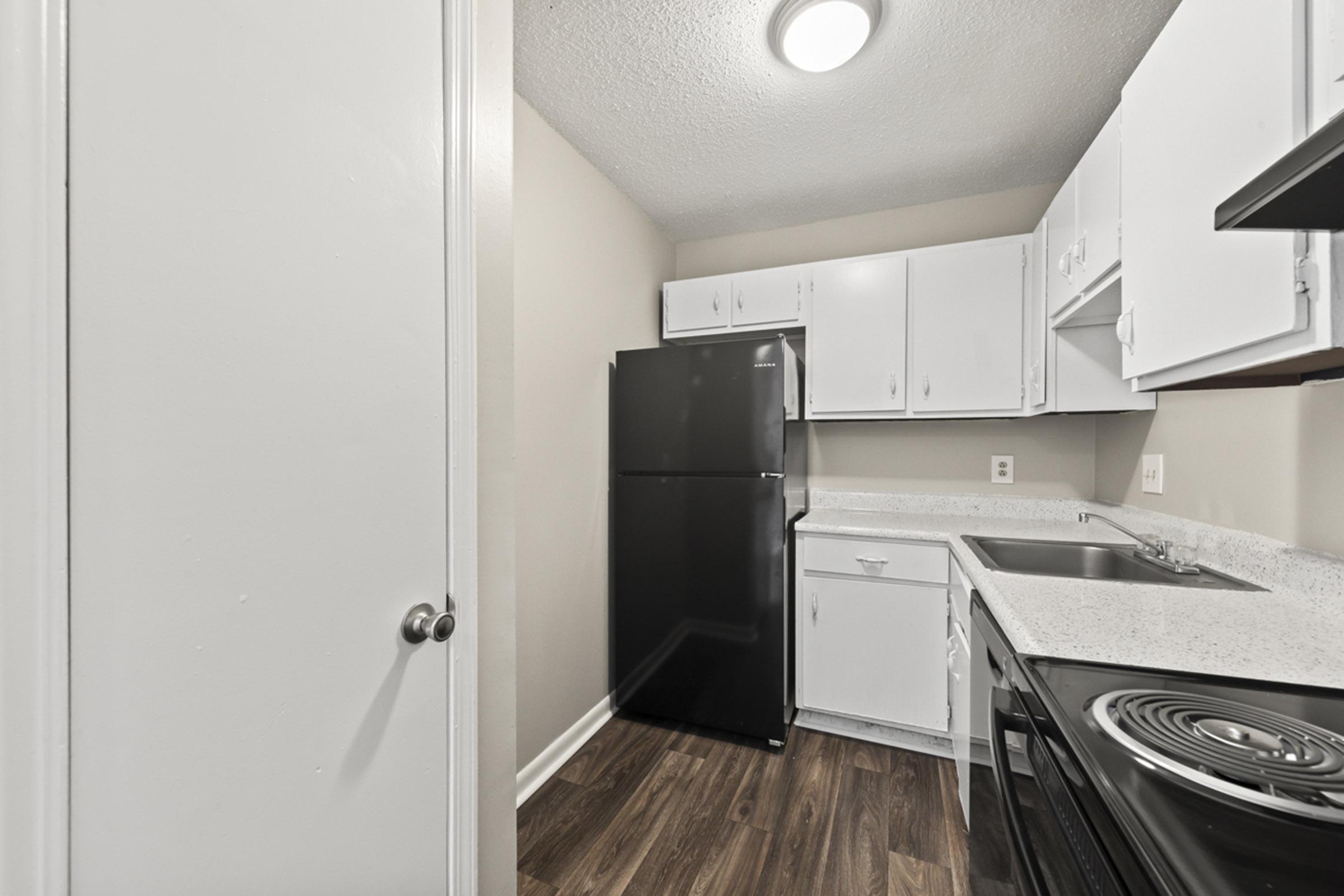 A small, modern kitchen featuring white cabinets, a black refrigerator, and a countertop with a sink and stove. The room has light walls and wooden flooring, creating a bright and tidy space. The layout is compact, with doors on the left and a light fixture above.