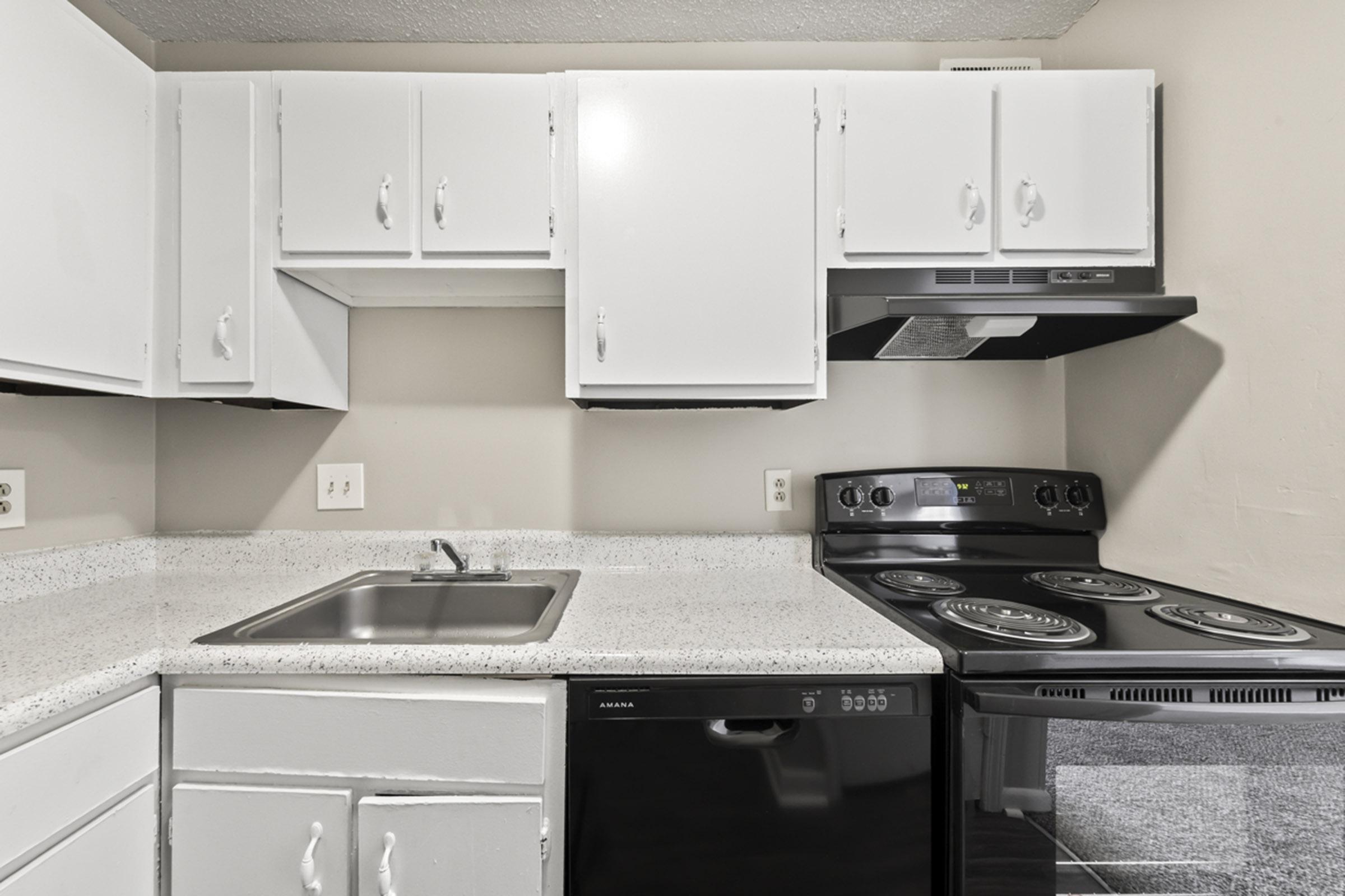 A modern kitchen featuring white cabinets, a stainless steel sink, a black dishwasher, and an electric stove with an overhead microwave. The countertop is made of speckled gray material, and the walls are painted a light color, creating a bright and functional cooking space.
