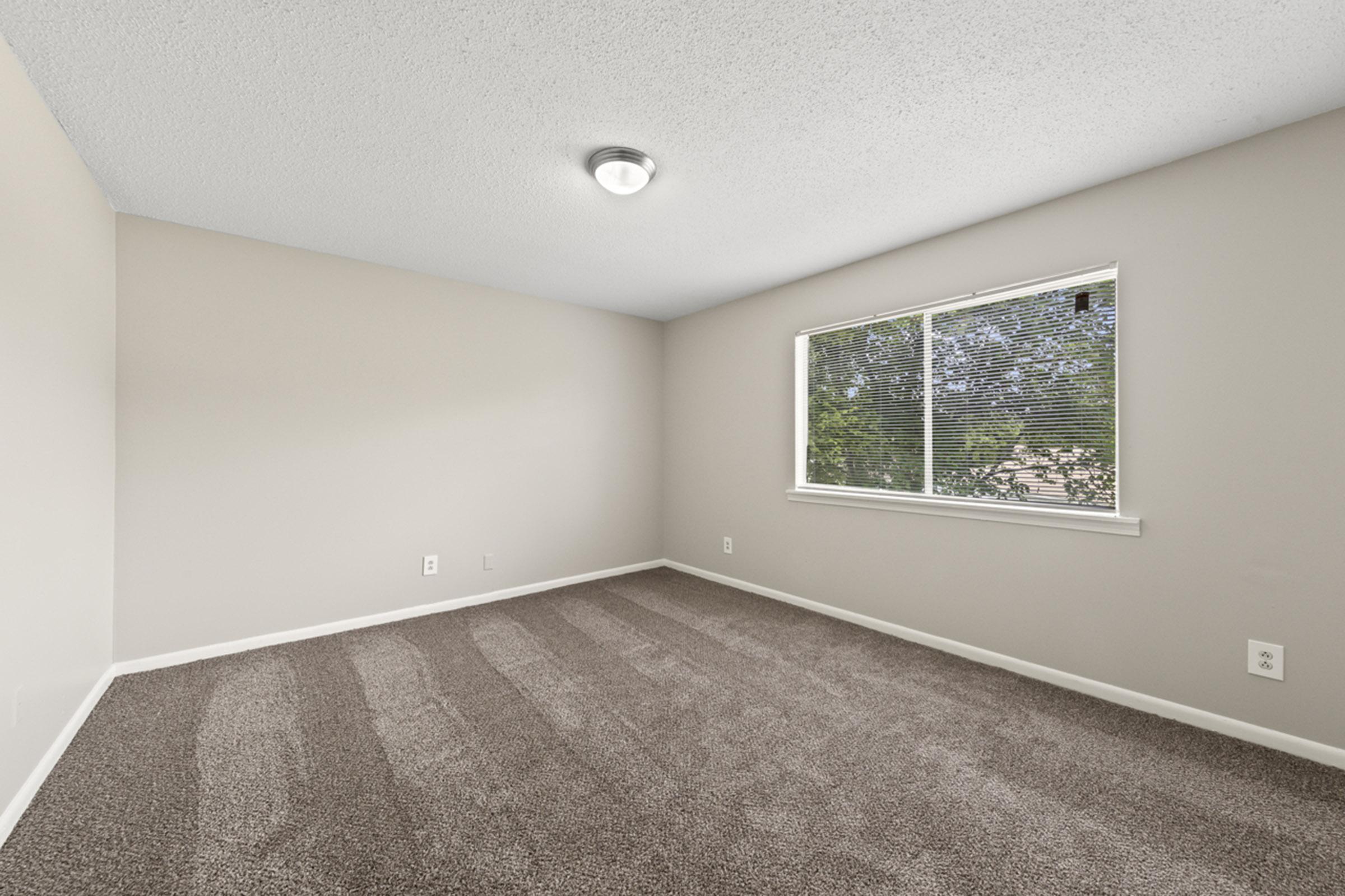 A vacant room with light beige walls and a textured ceiling. The floor is covered with light brown carpet, and there is a window on one wall allowing natural light to enter. The room appears spacious and unfurnished, offering a neutral backdrop for various design possibilities.