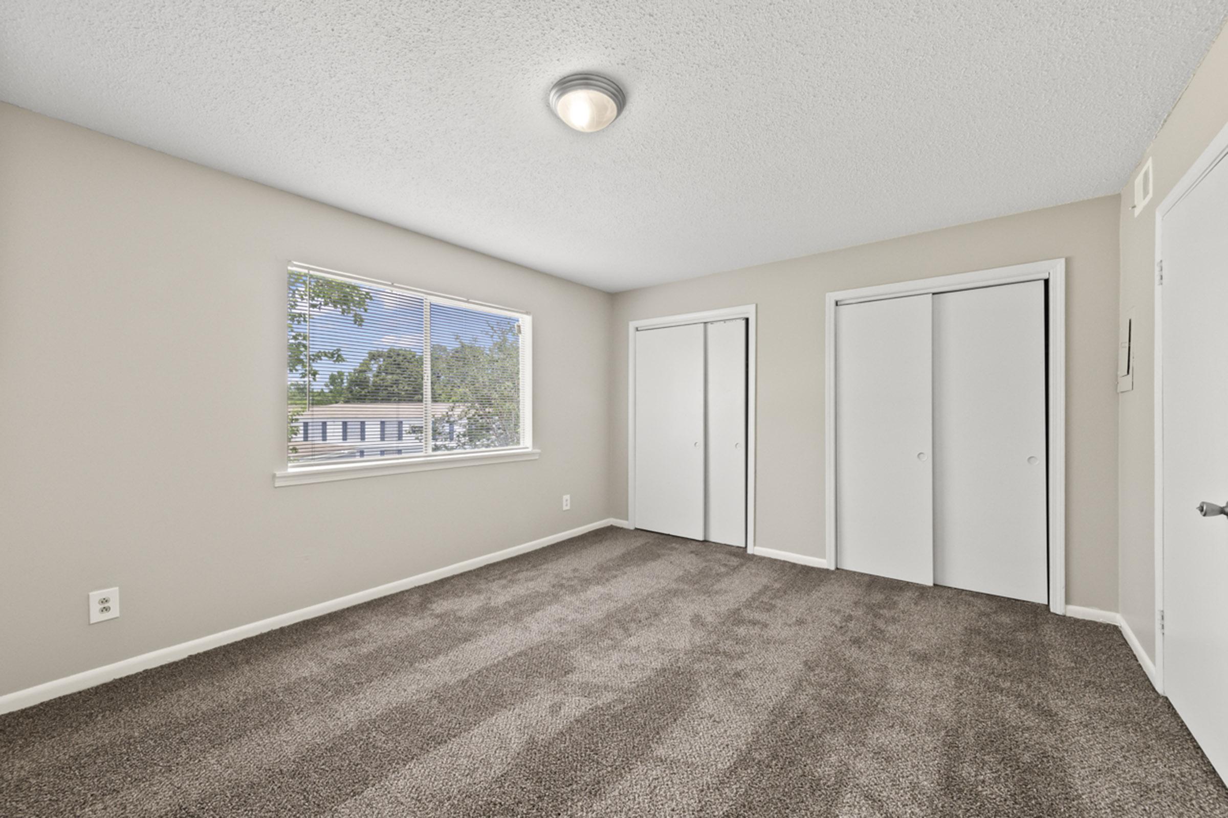 A well-lit room with light beige walls and carpeted flooring. It features a large window allowing natural light, a ceiling light fixture, and two white sliding door closets. The room is empty, providing a spacious and clean atmosphere.