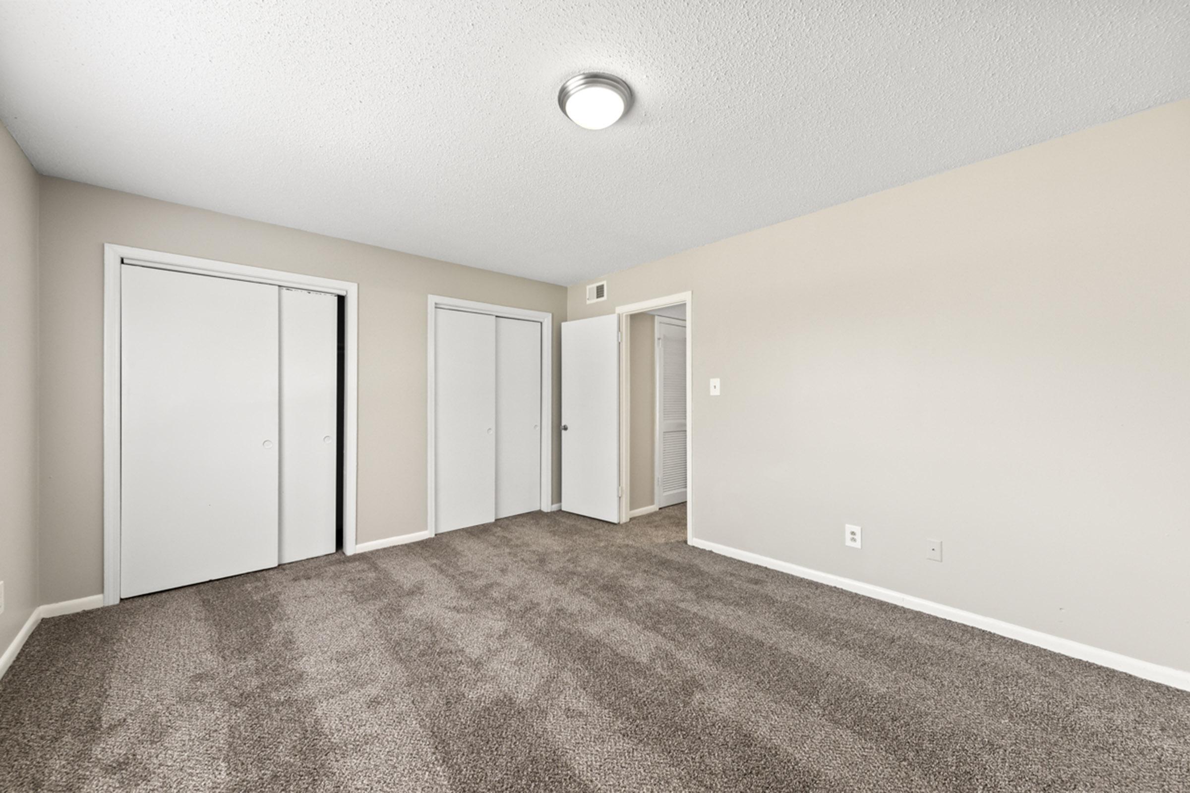 A spacious room featuring beige walls and plush carpet. There are two closed white closet doors on the left and a door leading to another space on the right. The ceiling has a round light fixture, and the overall atmosphere is bright and neutral, creating an inviting environment.