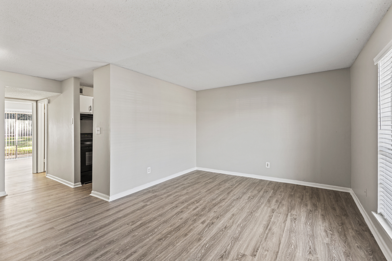 A spacious, vacant living room with light gray walls and wooden laminate flooring. Large windows allow natural light, and there's access to a patio or balcony area in the background. The kitchen is partially visible, featuring a black appliance setup. The overall ambiance is clean and inviting.