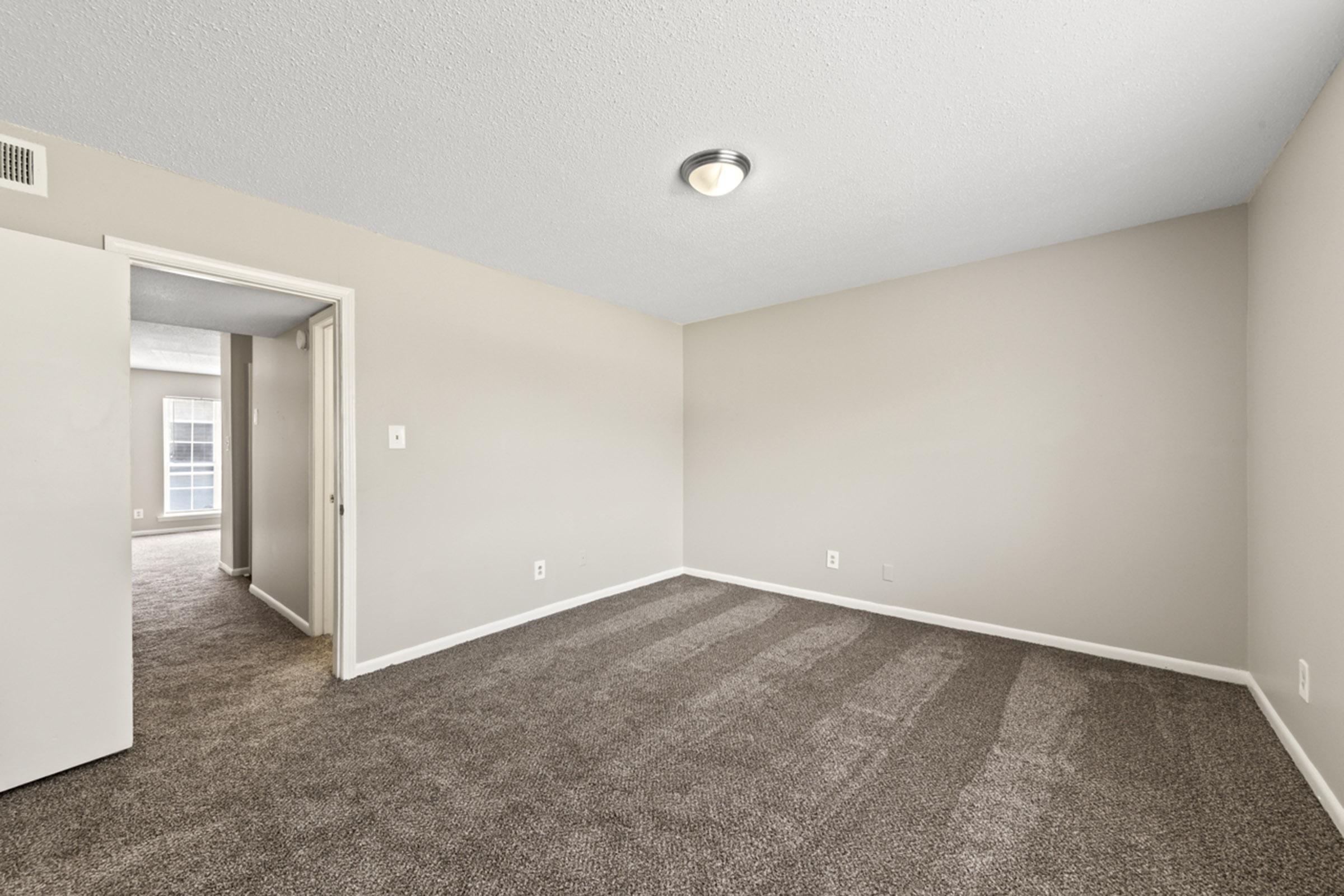Spacious empty room with beige walls and plush brown carpet, featuring a ceiling light. An open door leads to a hallway, and a large window provides natural light. The room appears bright and well-maintained, ideal for decorating or personalizing.