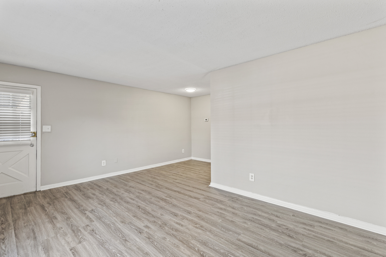Interior view of a spacious, empty room with light-colored walls and a wood-like vinyl floor. There is a single door with a window on the left side, and a ceiling light fixture above. The room has no furniture, creating a clean and minimalist look.