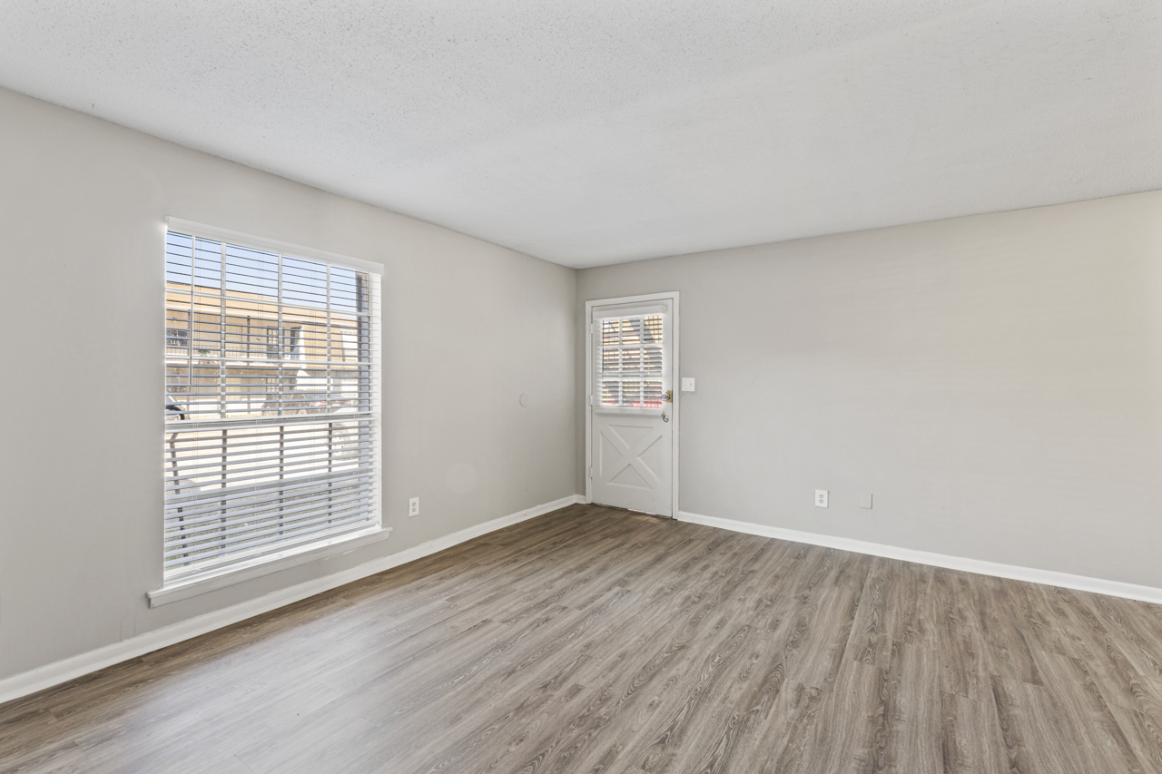 A vacant room with light gray walls and hardwood-style flooring. A window with white blinds allows natural light to enter, while a door with a glass panel is located on the right. The space appears bright and clean, suitable for various purposes such as a living area or office.