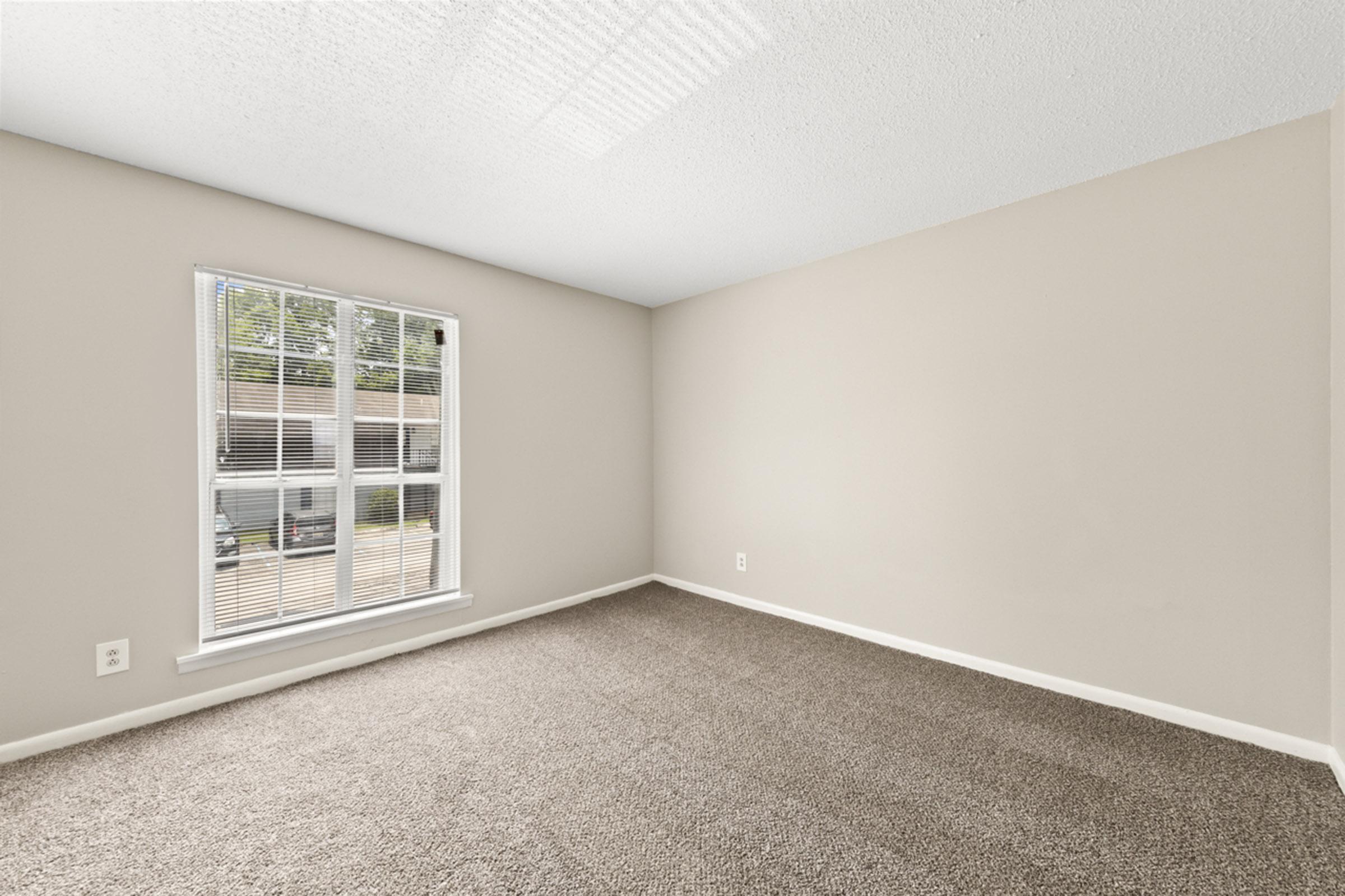 Interior of a vacant room with beige walls and carpeted flooring. A large window with blinds allows natural light to enter, illuminating the space. The room appears empty, offering a clean and neutral backdrop for various uses.
