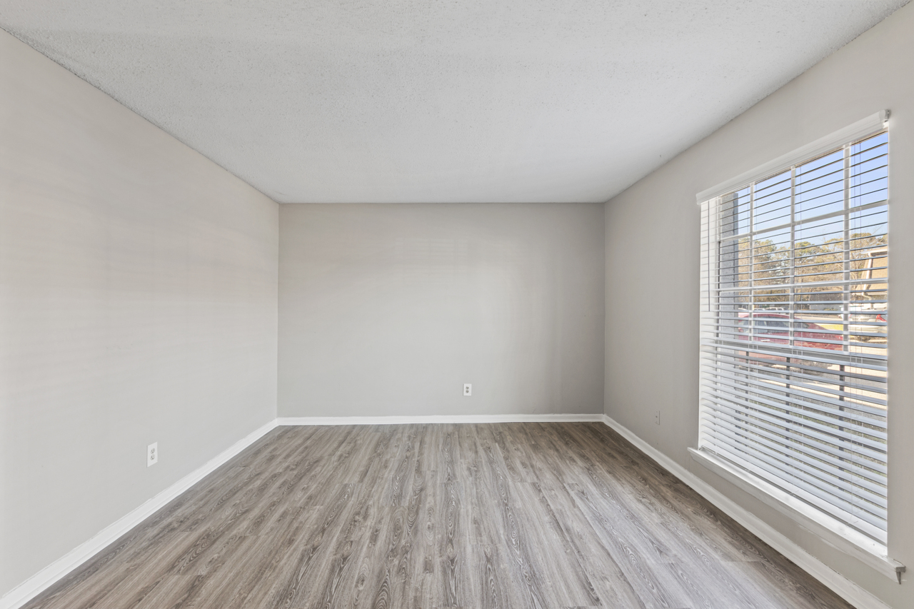 A spacious, empty room with light gray walls and laminate flooring. There is a window with white blinds letting in natural light. The room features clean lines and a simple, modern aesthetic, ideal for various potential furnishings or decorations.