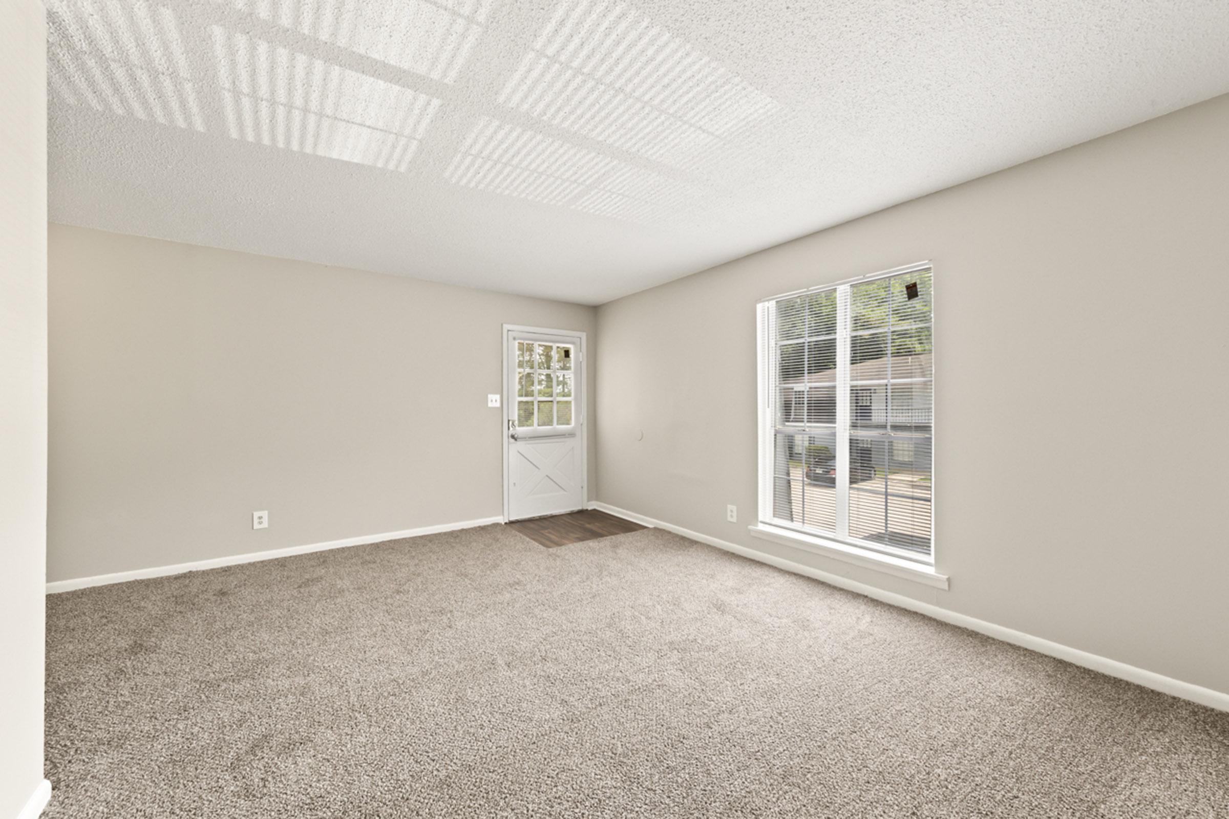 A spacious, empty living room featuring beige walls and carpet. A front door with a window is visible, along with a large window allowing natural light. The room has a simple design, highlighting its potential for furnishings and decor.