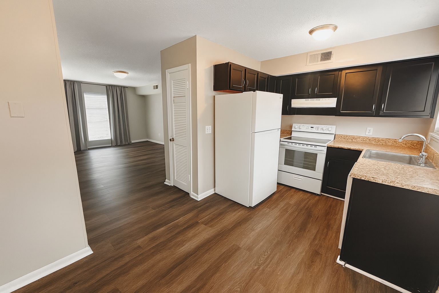 A spacious kitchen featuring dark cabinetry, a white refrigerator, and an oven with a stovetop. The kitchen has a granite countertop and is open to a well-lit living area with large windows and light-colored curtains. The flooring is a warm wood tone, creating a cozy atmosphere.