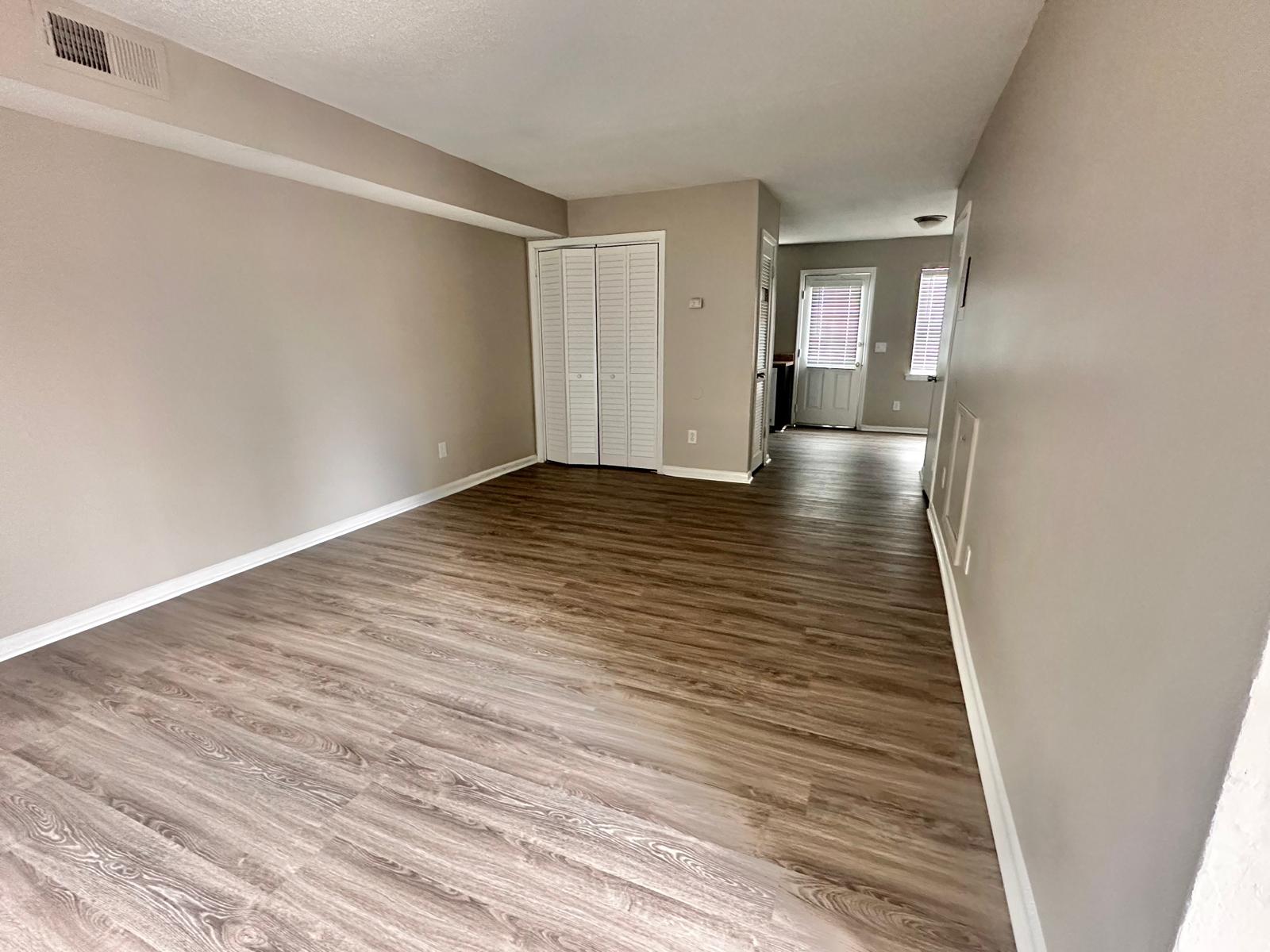 Empty room with light brown hardwood flooring, painted light walls, and a closet with sliding doors on the left. A doorway leads to another room at the far end, and a door to the outside is visible on the right side. Natural light enters through the windows.