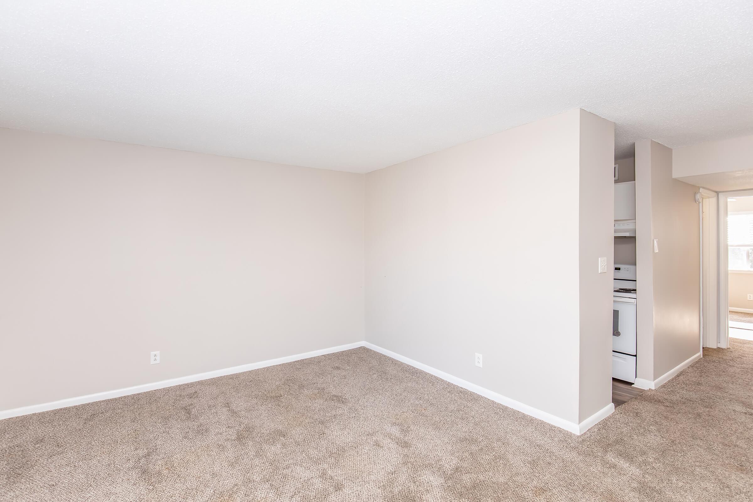 A spacious, empty room with light beige walls and carpet. There is an opening leading to a kitchen area, visible in the background. The room is well-lit and features clean lines, creating an inviting atmosphere for potential decoration or furnishing.