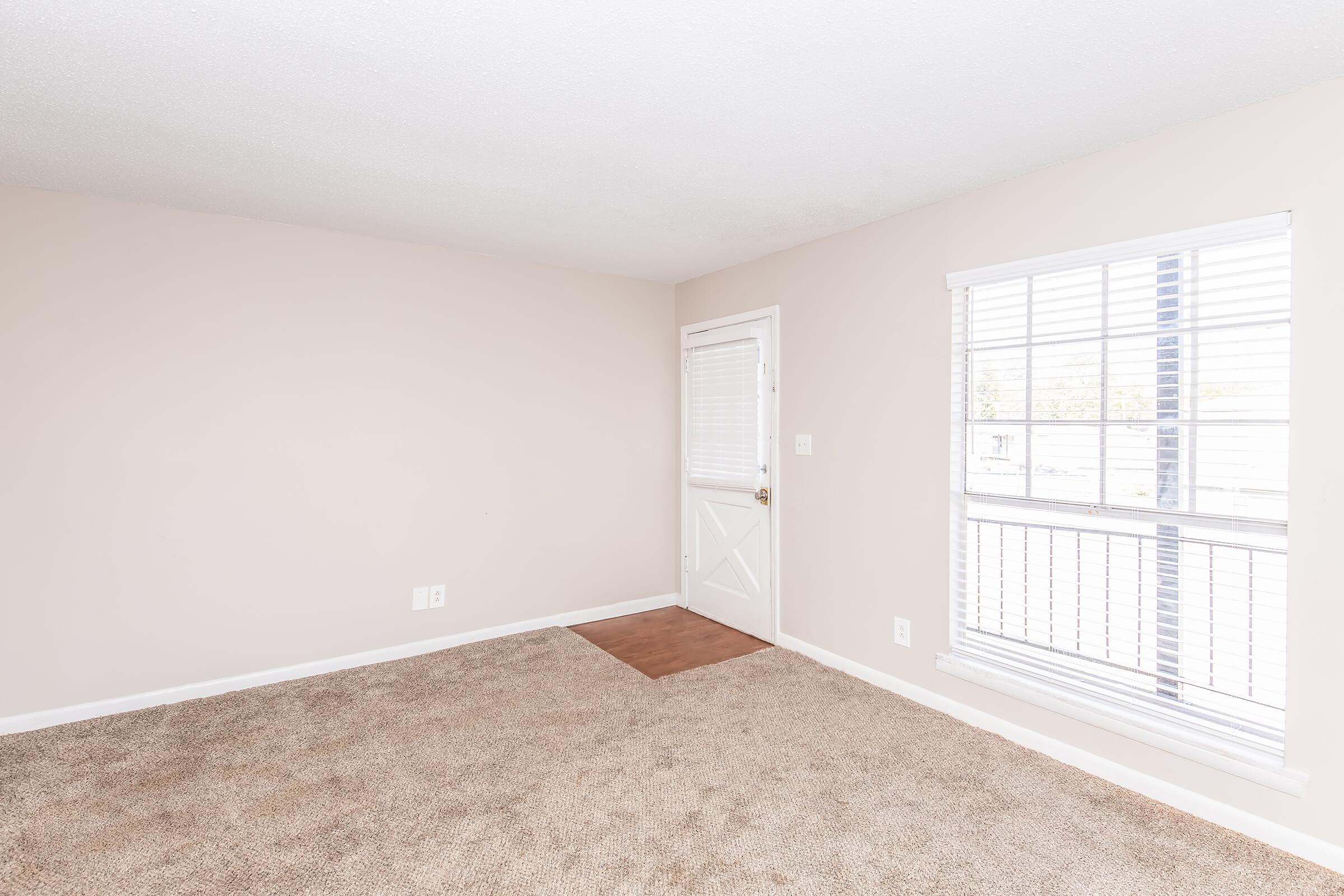 A light-colored room featuring beige walls and carpet, with a white door on the right side leading outside. There are two windows with white blinds allowing natural light to enter. The space appears empty and clean, offering a neutral backdrop for various furnishings.