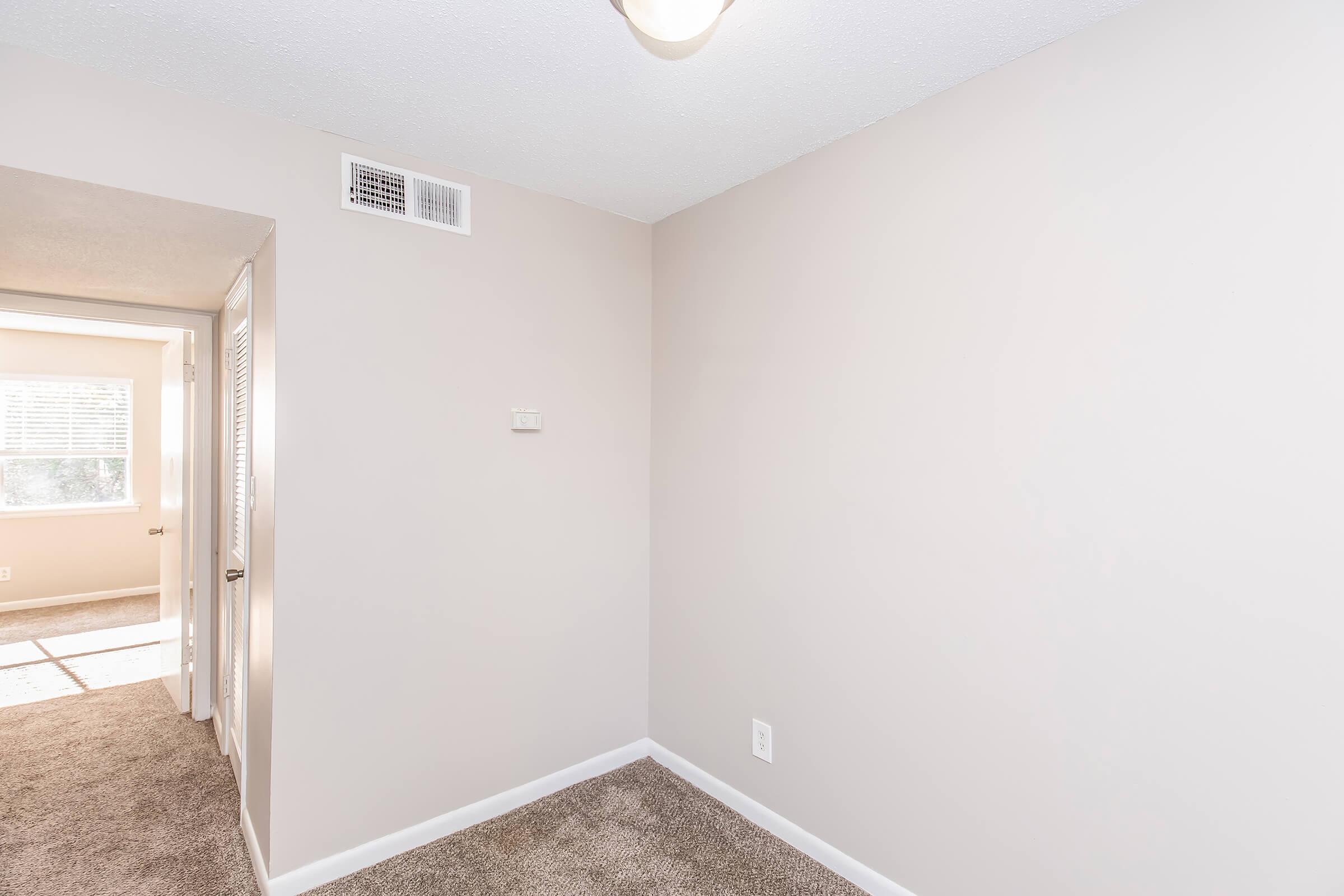 A well-lit, neutral-colored room with a carpeted floor and a light fixture on the ceiling. One wall is bare, and there's a door on the left leading to another room. A window is visible further down the hallway, providing natural light. The overall atmosphere is clean and spacious.