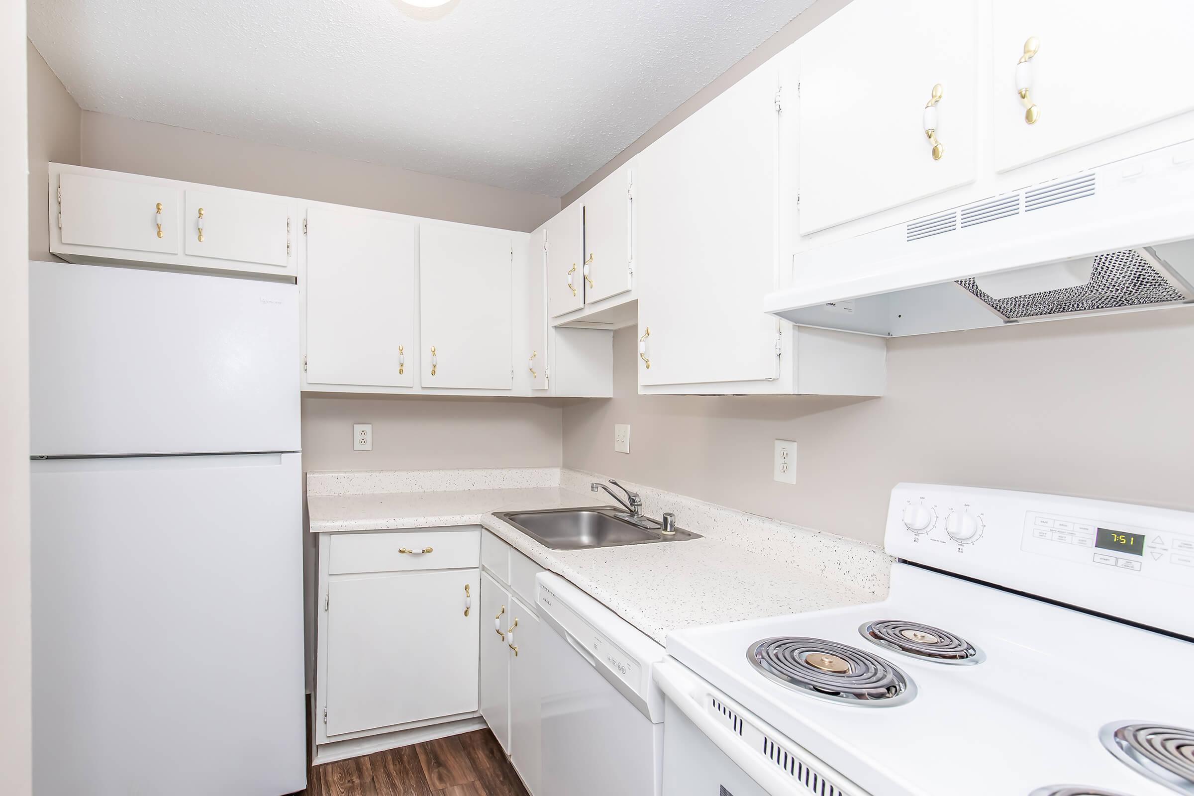 A clean, modern kitchen featuring white cabinets, a refrigerator, a dishwasher, and a stove with an oven. The countertop is light-colored and the walls are a neutral shade, creating a bright and inviting space. The floor is a wood-like finish, adding warmth to the overall design.