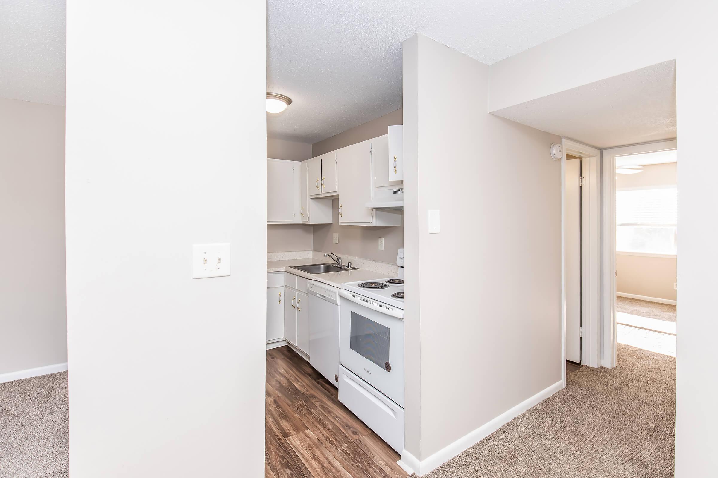 A small, modern kitchen featuring white cabinetry, a stove, oven, and sink. The walls are painted in a neutral color, and the floor is covered with laminate flooring. There is a doorway leading to another room, and the space is well-lit with natural light.