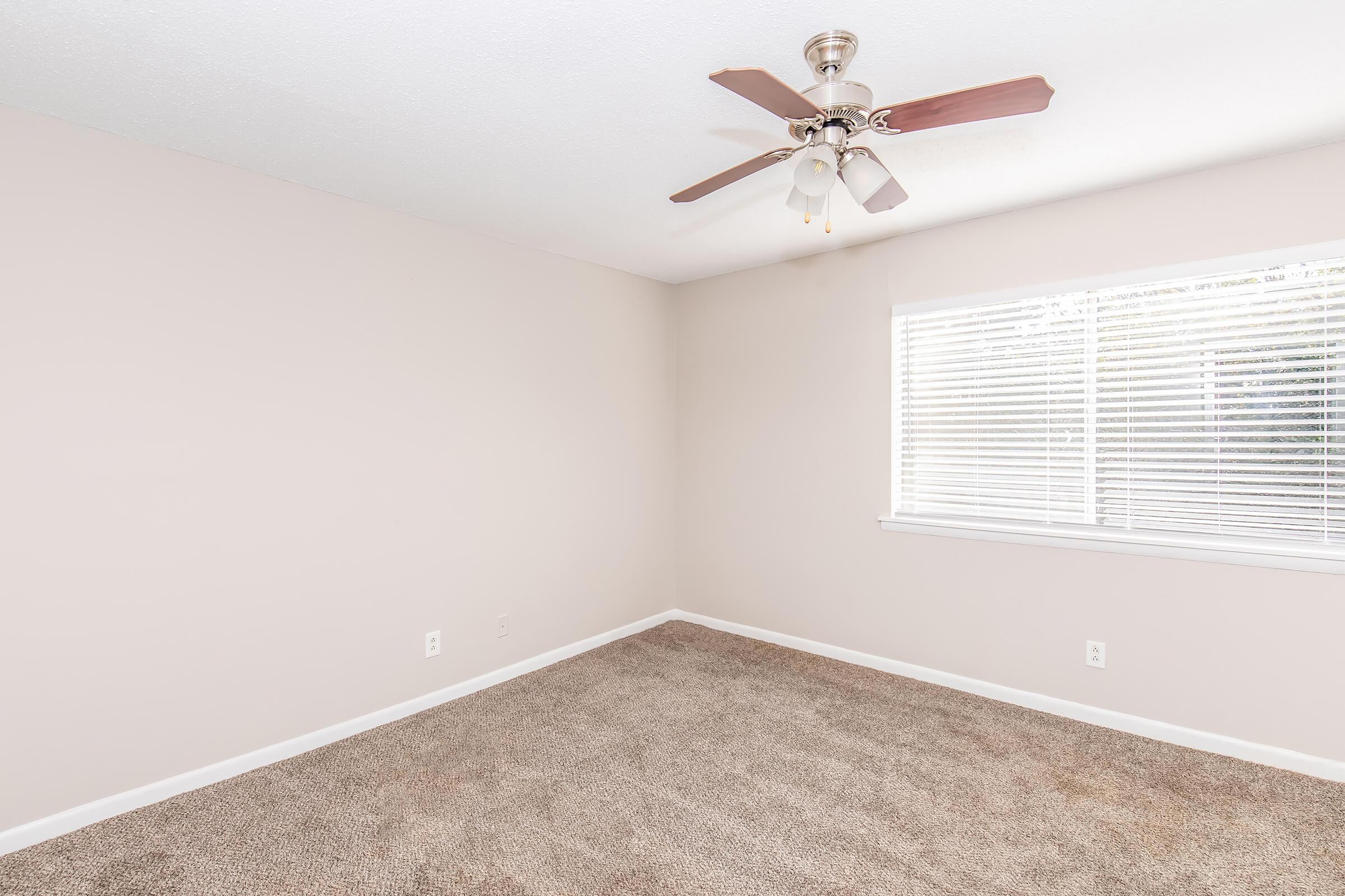 A bare room with light beige walls, a ceiling fan with wooden blades, and a window covered by white blinds. The floor is carpeted in a light color, and the room is empty, featuring only a few electrical outlets on the walls. The overall ambiance is bright and neutral.