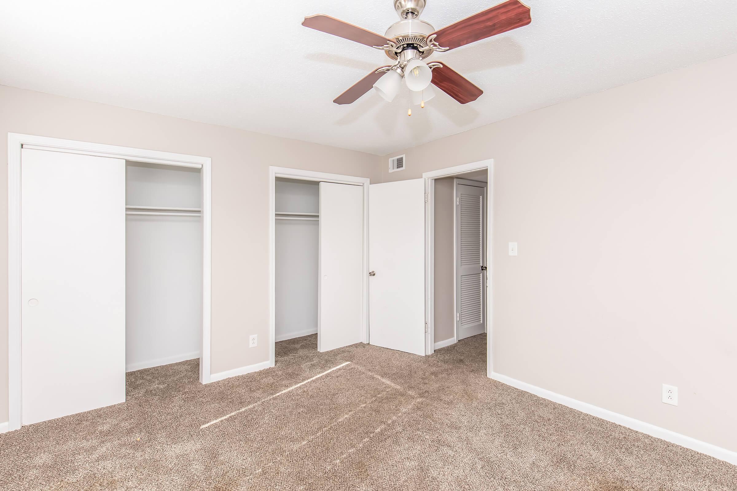 A neutral-colored bedroom featuring light beige walls and a ceiling fan with wooden blades. There are two closet doors on the left and a white door leading to another room on the right. The floor is covered in light brown carpet, and natural light is visible through the doorway.