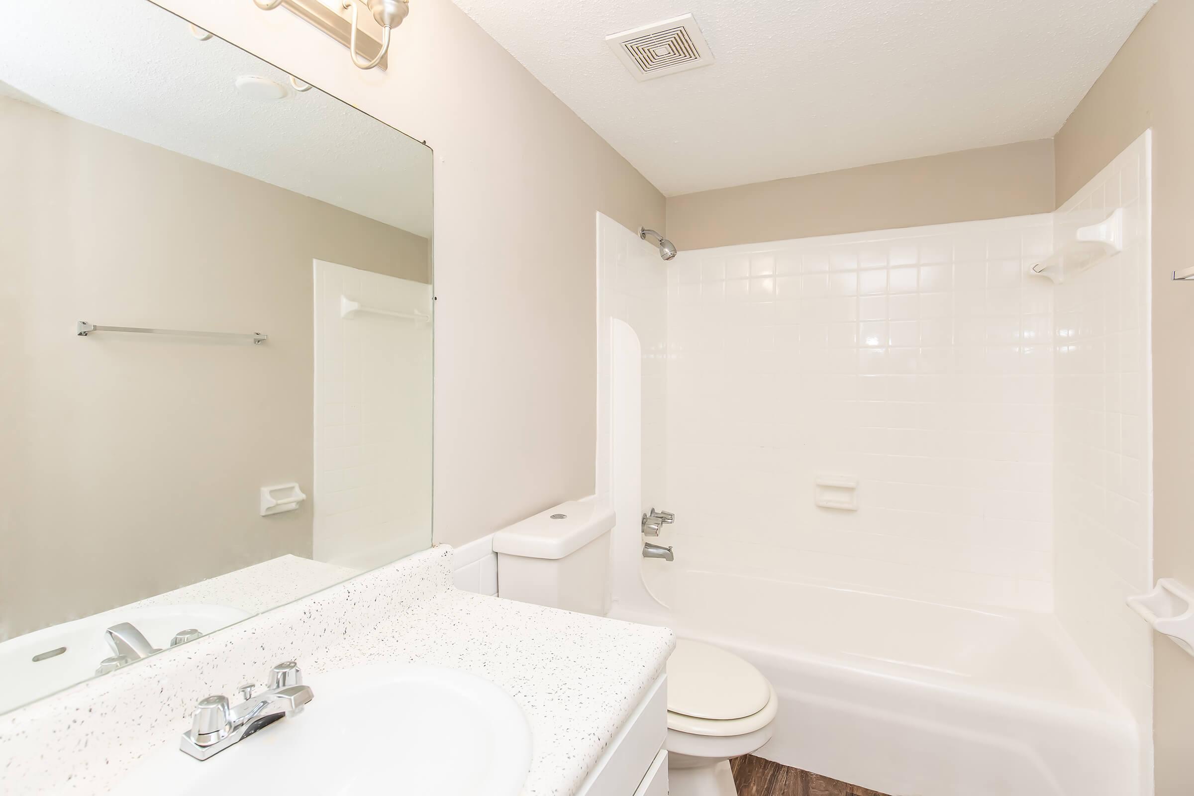 A clean, modern bathroom featuring a white bathtub with a shower, a toilet, and a sink with a speckled countertop. A large mirror reflects the space, and neutral-colored walls enhance the bright, airy feel. Minimalistic design with essential fixtures and good lighting.